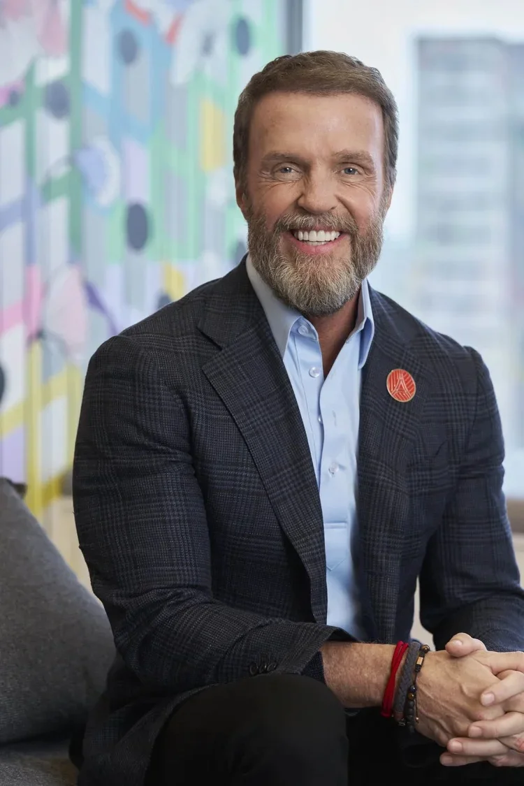 Smiling middle-aged man with a beard and short brown hair, wearing a dark plaid blazer over a light blue shirt, sitting in an office with colorful artwork in the background.
