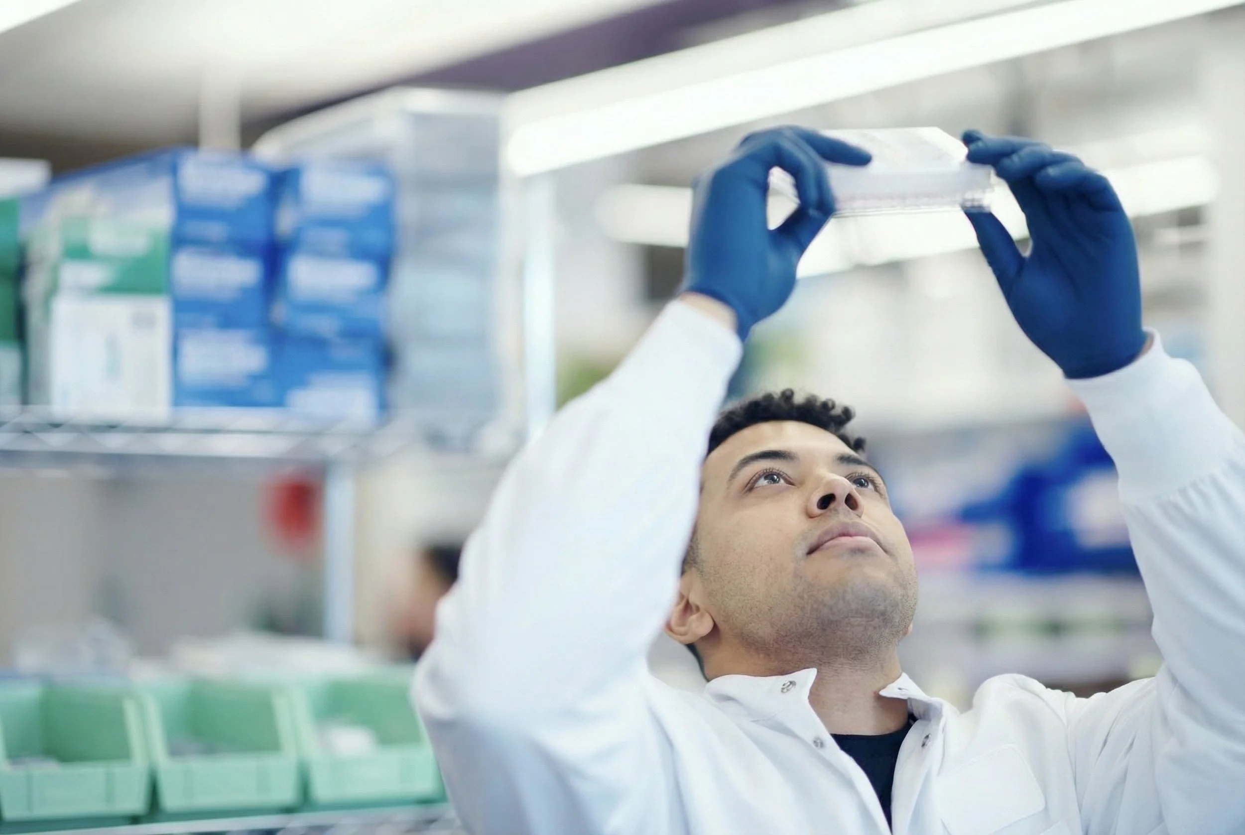 A scientist wearing a white lab coat and blue gloves is handling a transparent plastic container in a laboratory setting with shelves of laboratory supplies in the background.
