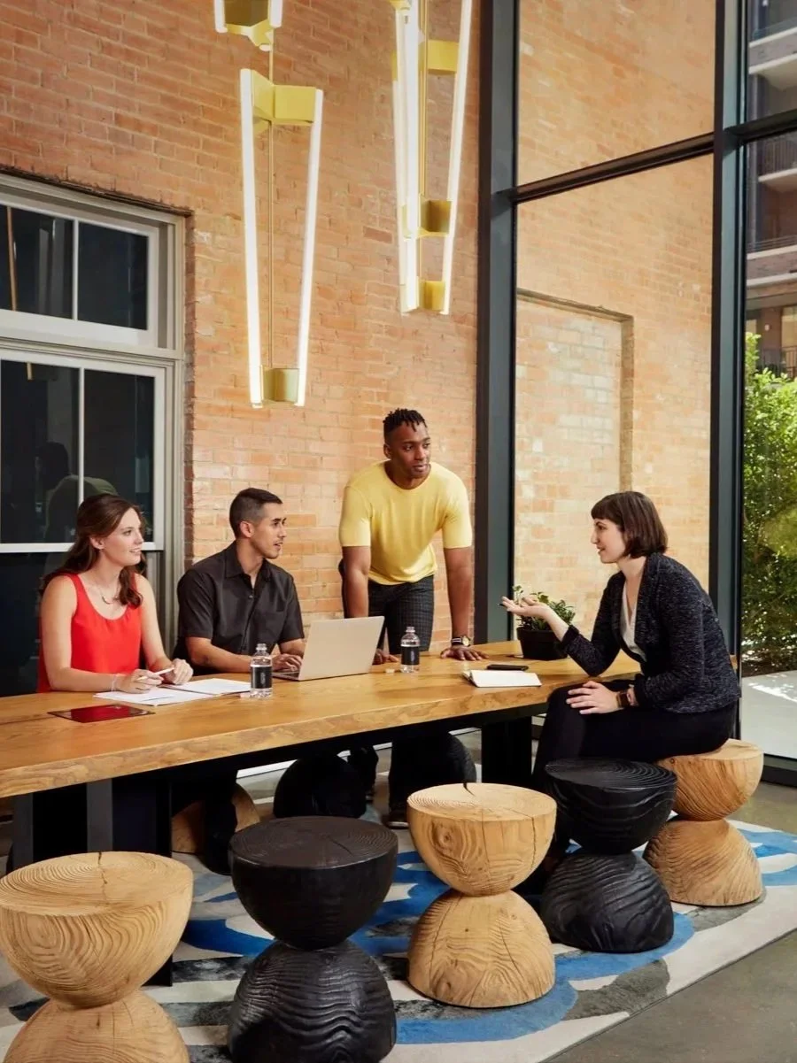 Four people in a meeting room with a brick wall and large window. Three seated on one side of a wooden table, one standing in front of a laptop. The room has hanging light fixtures and stools around the table.