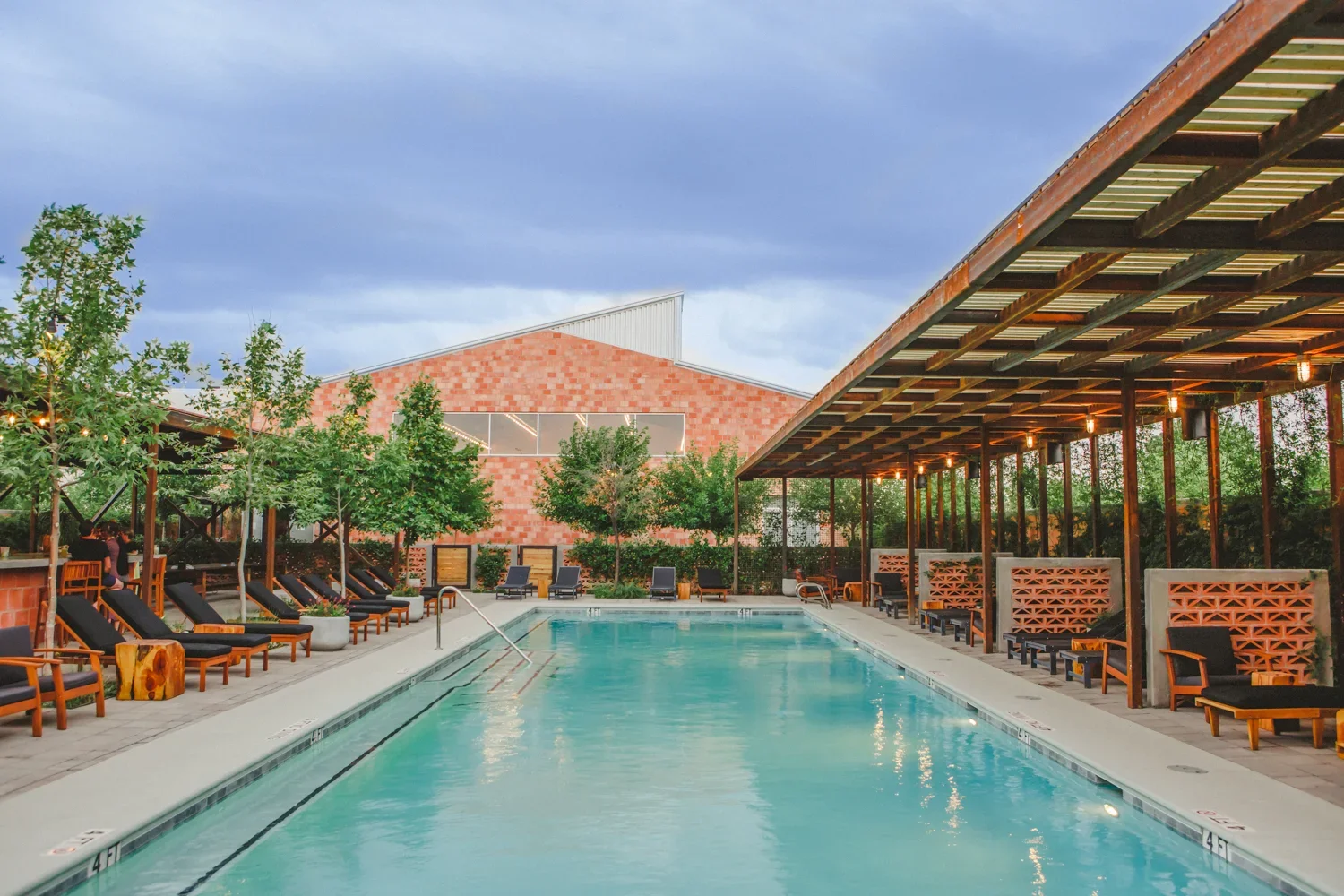 Outdoor swimming pool surrounded by lounge chairs and shaded seating areas, with trees and a brick building in the background under a cloudy sky.