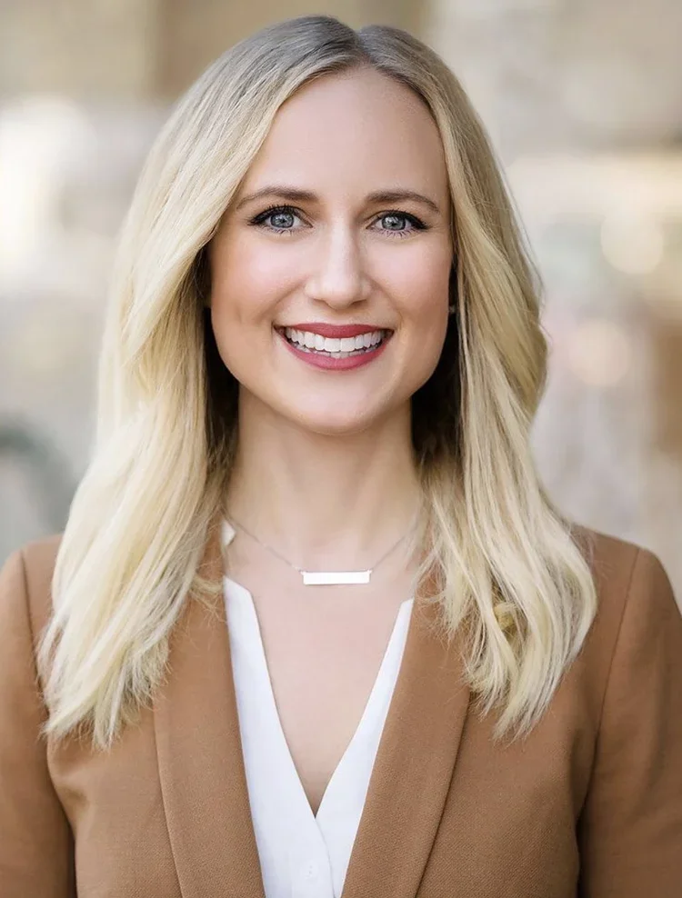 A smiling woman with long blonde hair, wearing a beige blazer and a white top, standing outdoors.