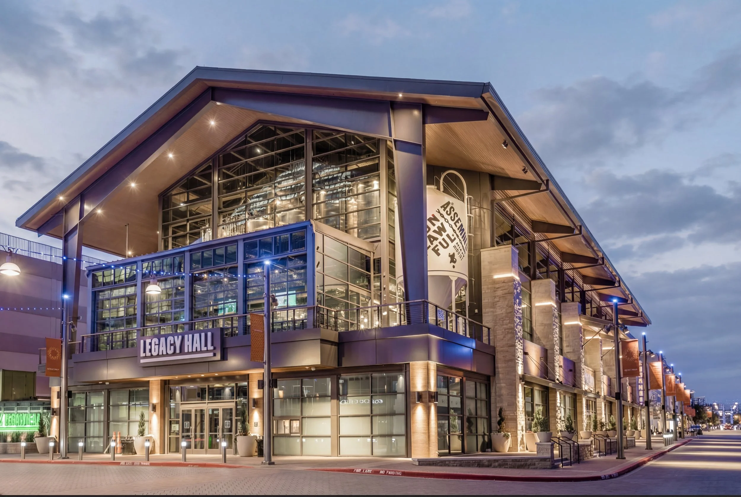 Exterior view of Legacy Hall, a modern multi-story building with large glass windows, illuminated at dusk, featuring a sign with its name on the front, and surrounding streetlights and sidewalk.