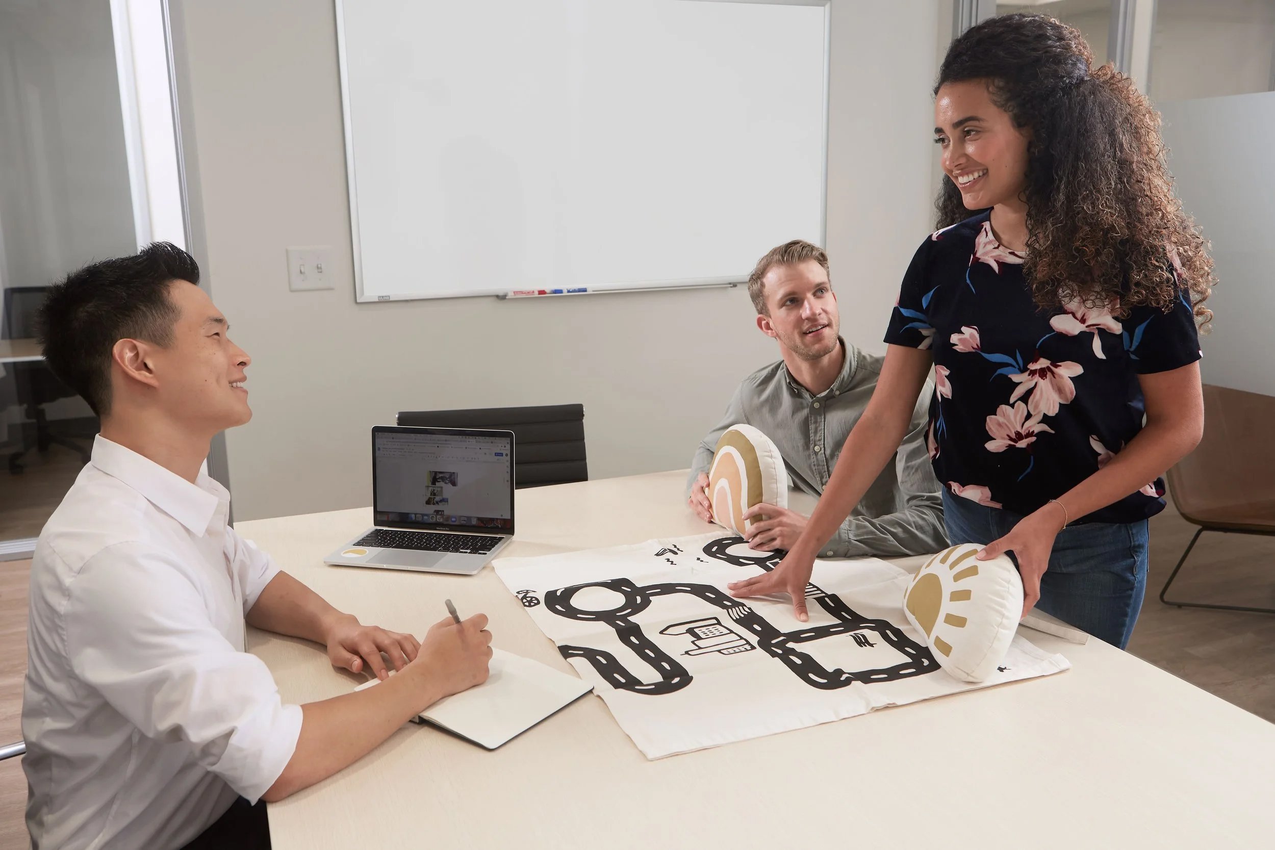 Two men and a woman in an office meeting, with a large paper drawing of a road map and plush objects on the table.