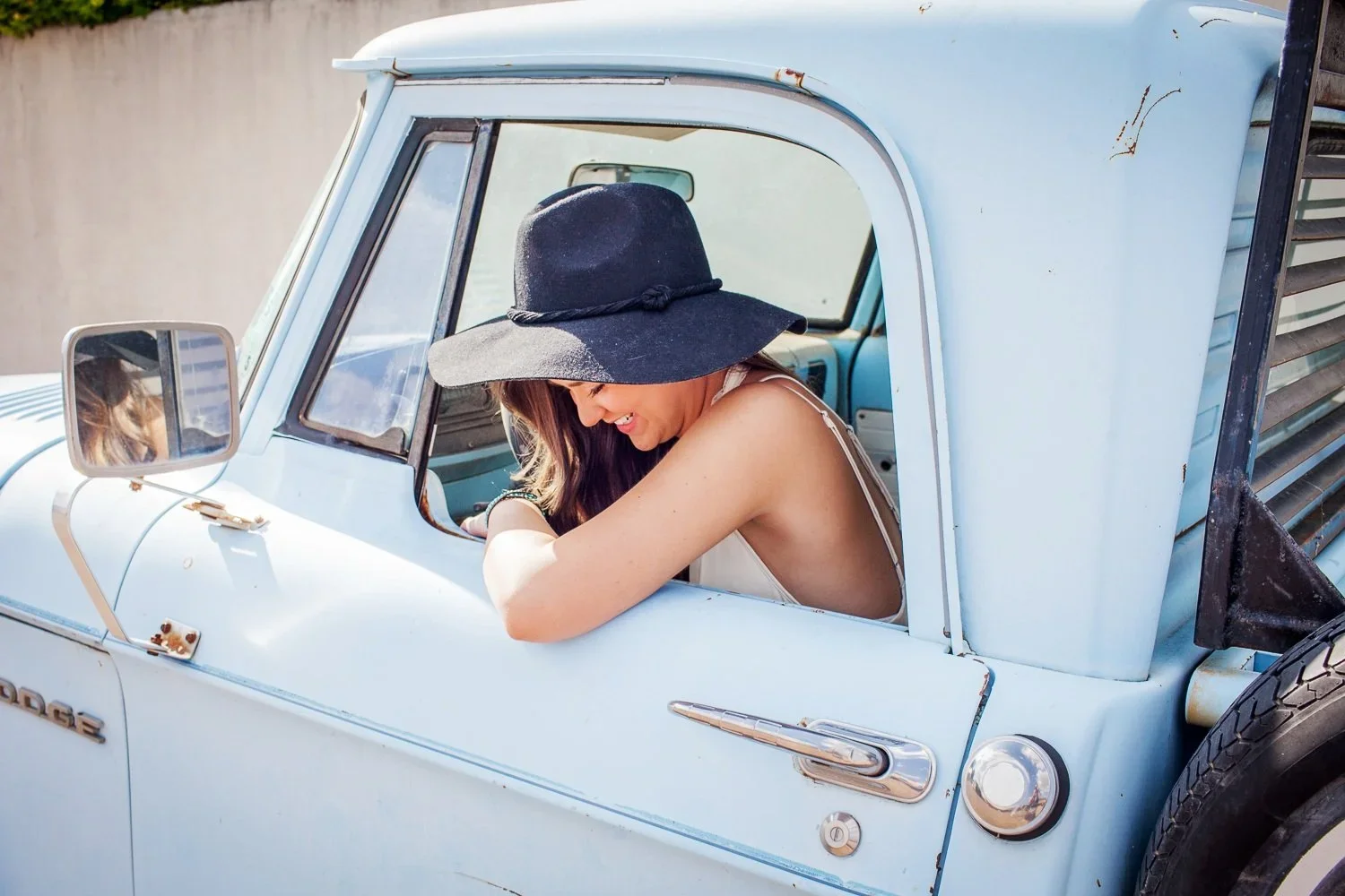 A woman wearing a black wide-brimmed hat and a sleeveless top leaning out of a light blue vintage truck, smiling and looking down.