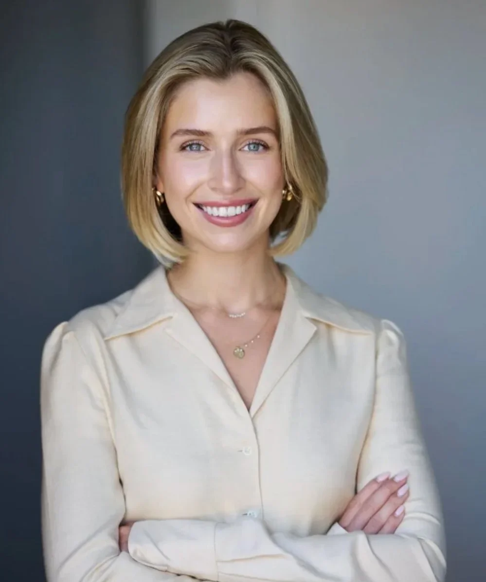 A smiling woman with blonde hair, wearing a beige shirt, gold hoop earrings, and layered necklaces, crossing her arms against a blurred background.