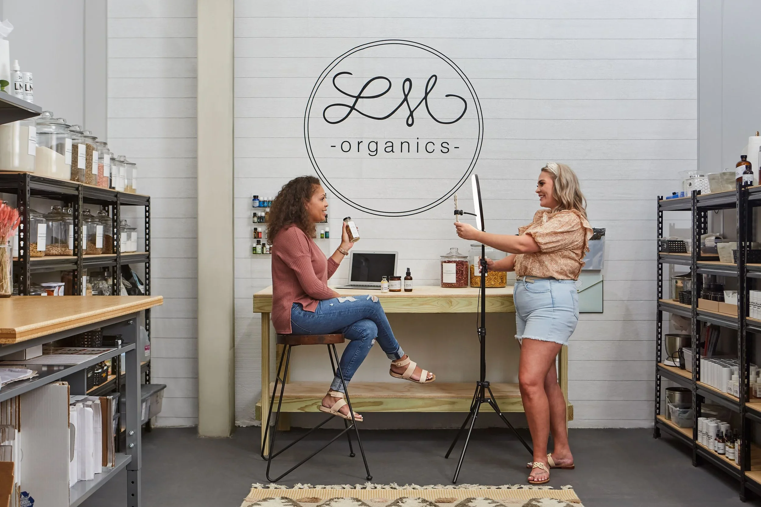 Two women in a store called LS Organics: one woman is sitting on a stool and holding a product, the other is standing with a camera, recording her. The store has shelves with jars and bottles, and a large logo on the wall that says LS Organics.