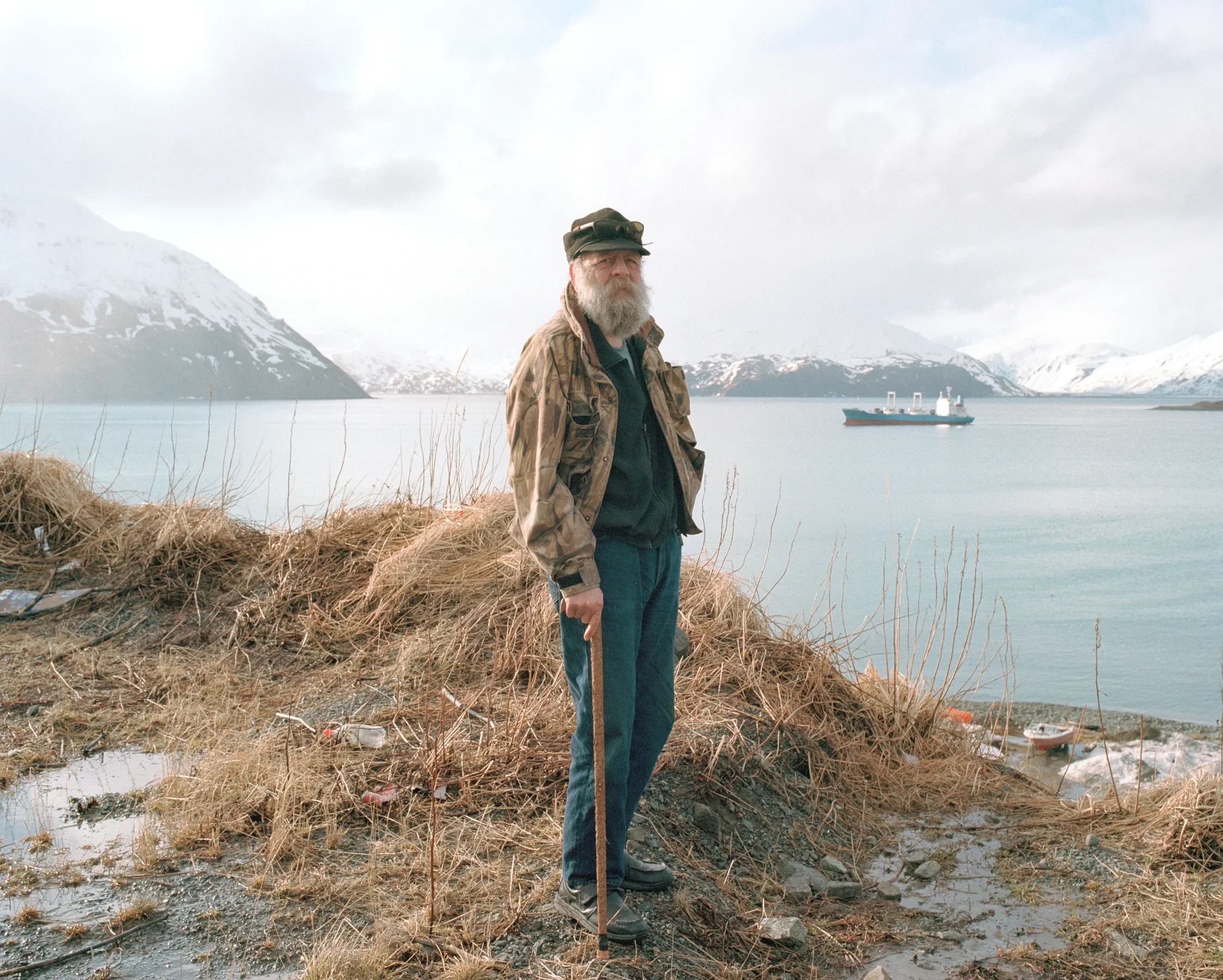 Older man with a beard wearing a camouflage jacket, blue jeans, and sunglasses on his hat, standing on a grassy shore with a walking stick, overlooking a fjord with mountains, a boat, and a cloudy sky.