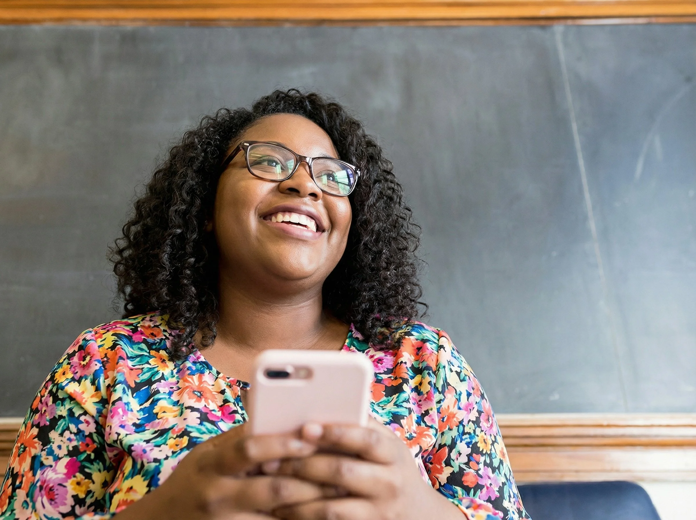A woman with curly hair and glasses smiling while looking at her phone, sitting in front of a chalkboard.