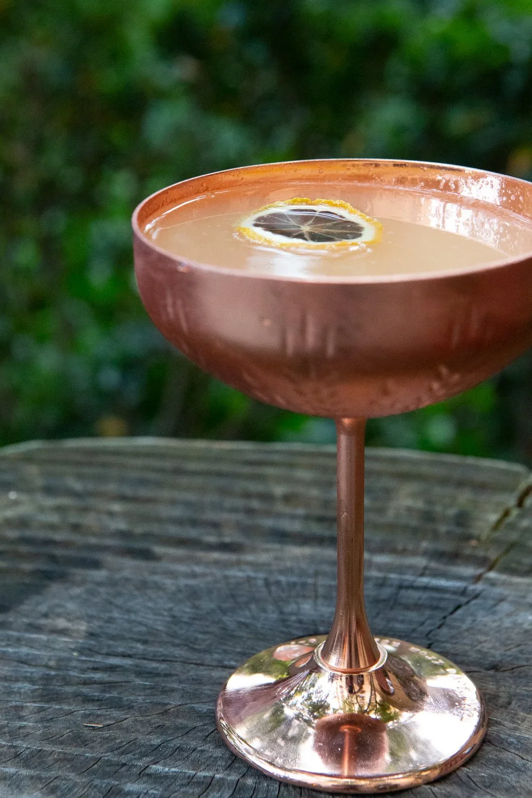 A copper cocktail glass on a wooden surface, containing a drink garnished with a lemon and lime slice, with a blurry green background.