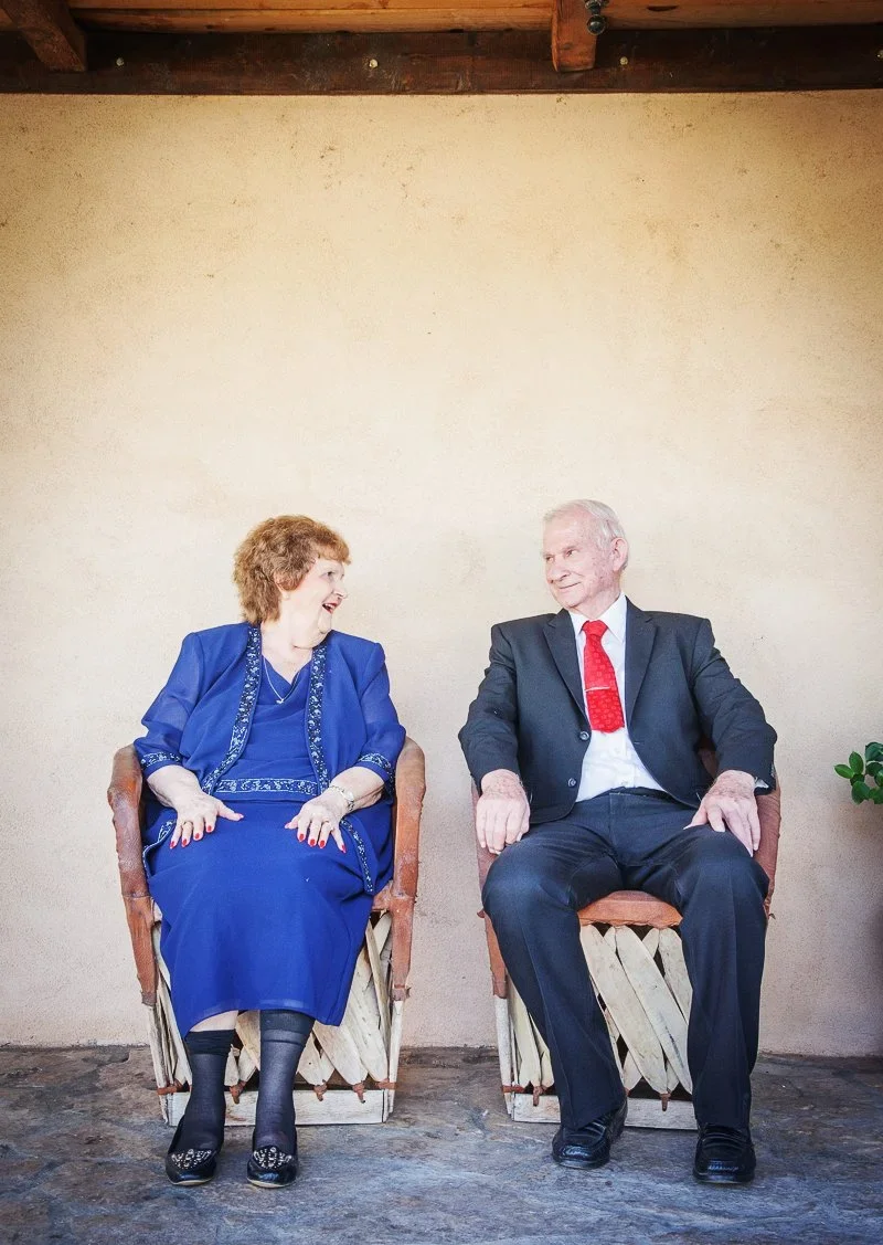 An elderly woman and man sitting in chairs against a beige wall, smiling at each other during a conversation.