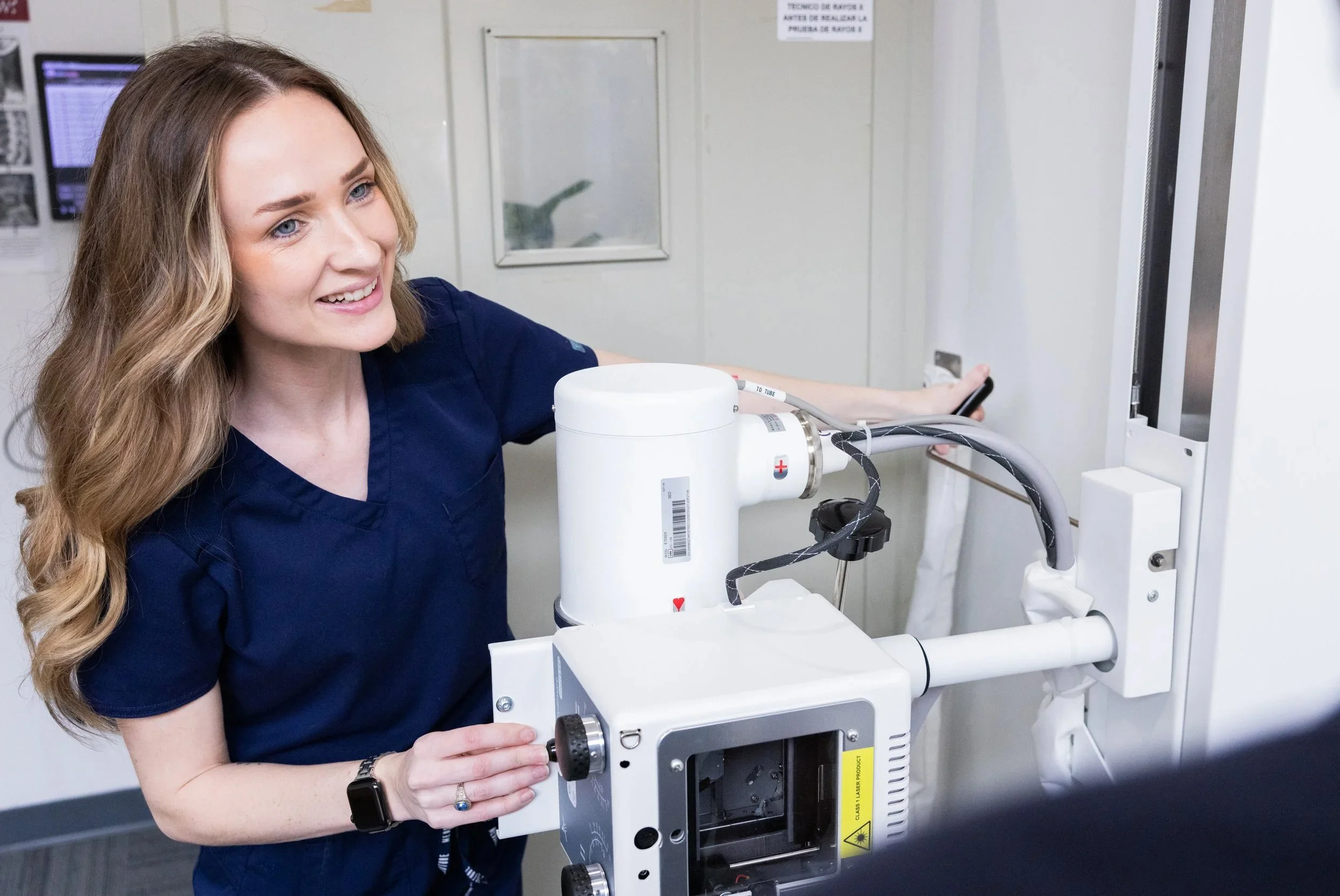 A female healthcare worker with long wavy hair wearing navy scrubs smiling while operating a medical imaging device in a hospital or clinic setting.