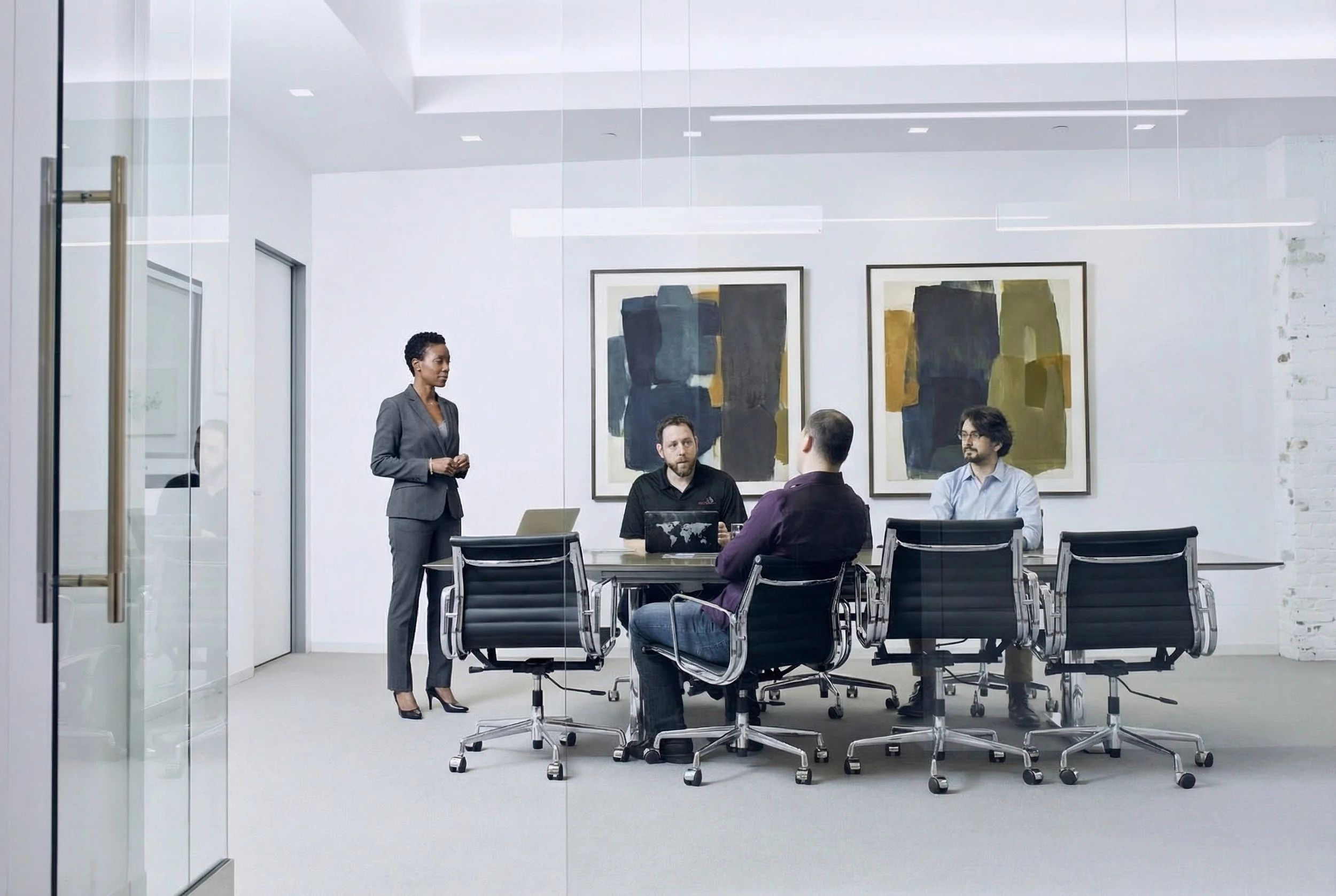 A businesswoman in a gray suit standing and speaking to three seated men in a modern conference room with abstract artwork on the wall.
