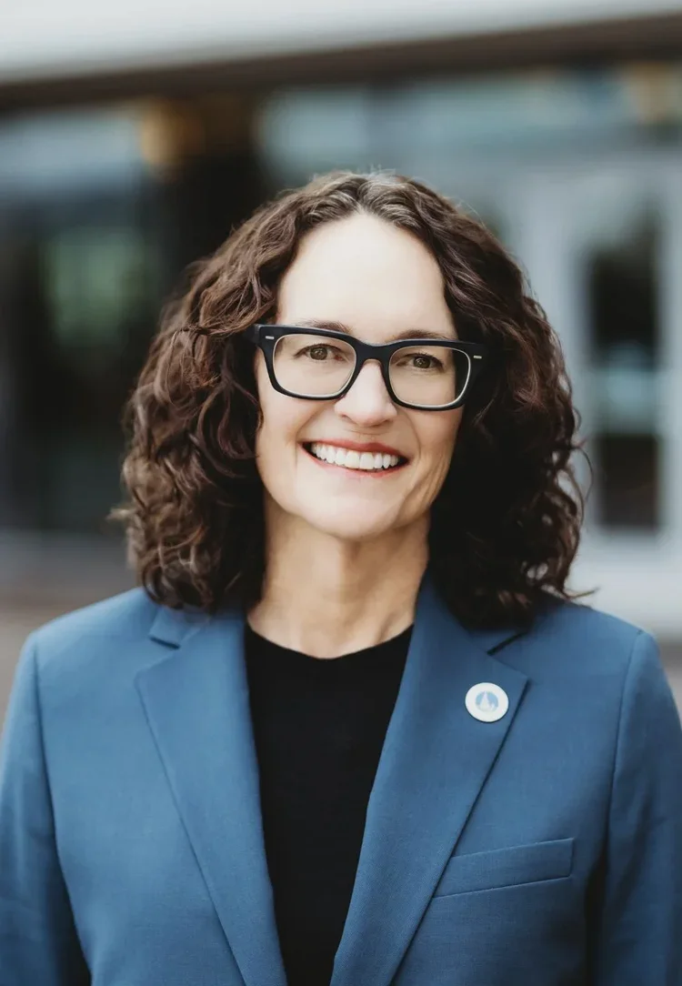 A smiling woman with curly brown hair, black glasses, a blue blazer, and a black top standing outdoors in front of a blurred background.