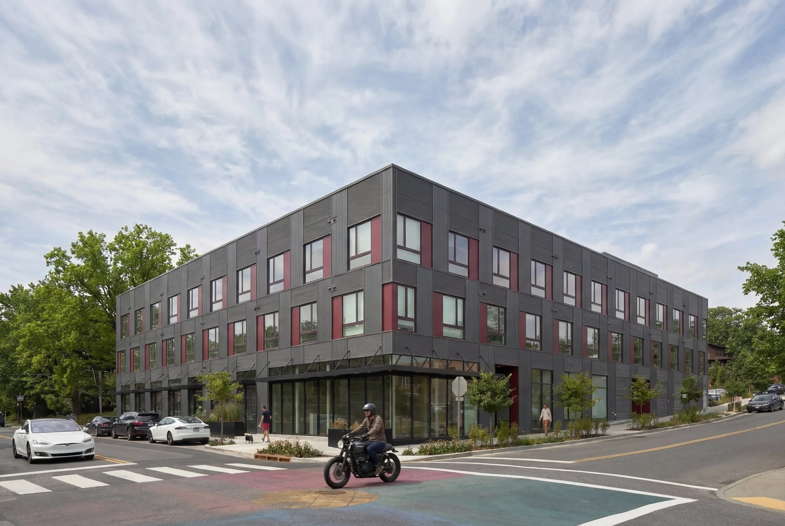 Modern black commercial building with large windows on a city street corner, cars and pedestrians, and a motorcyclist passing by, under a partly cloudy sky.