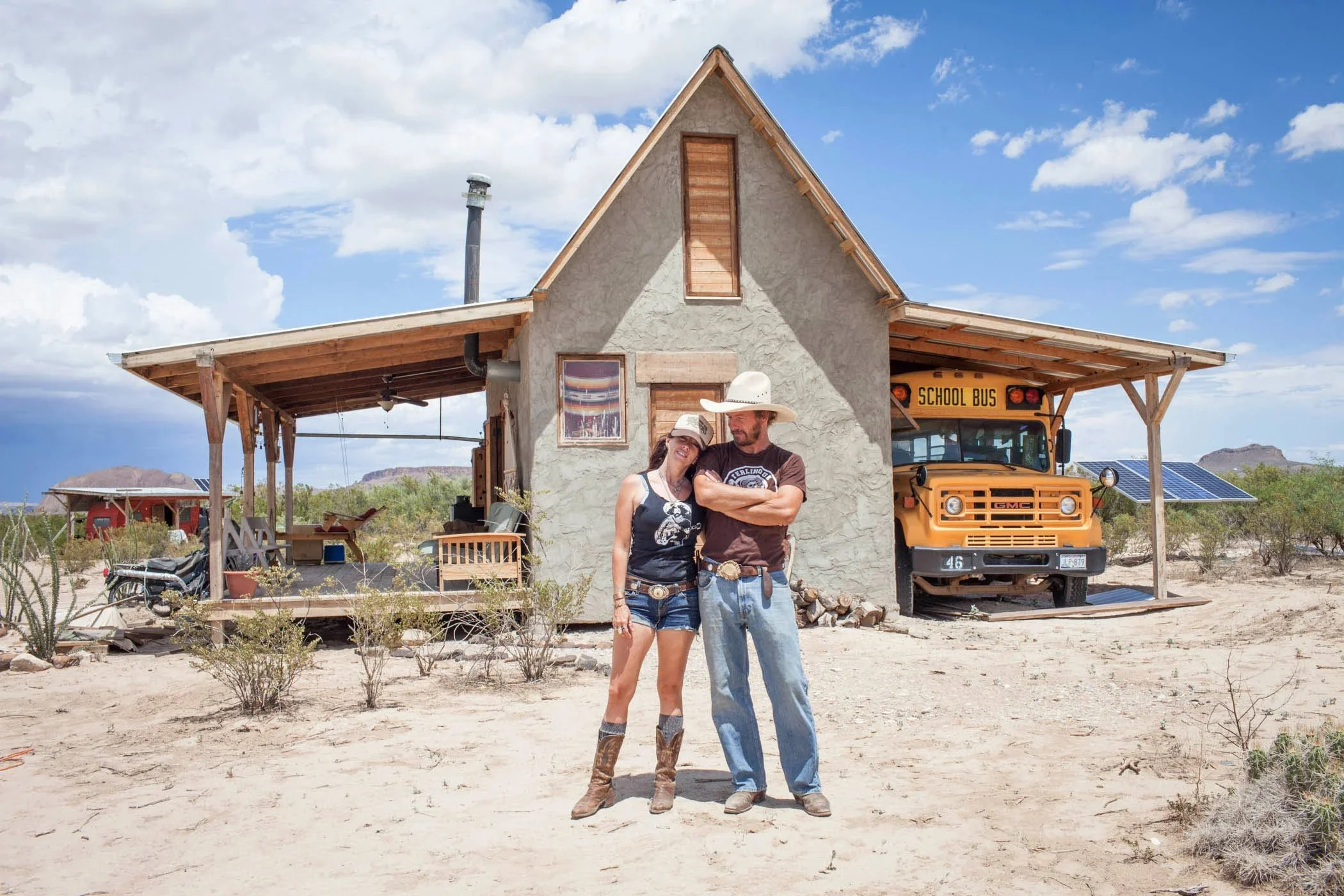 A couple dressed in cowboy attire standing in front of a small house with a vintage school bus parked inside a covered area. Solar panels are visible on another building in the background in a desert landscape.