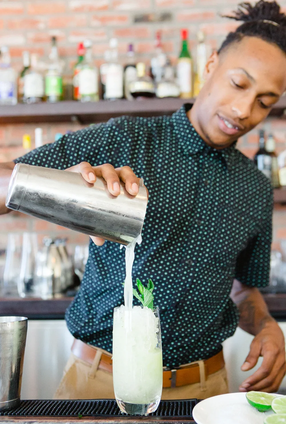 Bartender pouring a cocktail into a glass, garnished with mint leaves and lime, with liquor bottles on shelves in the background.