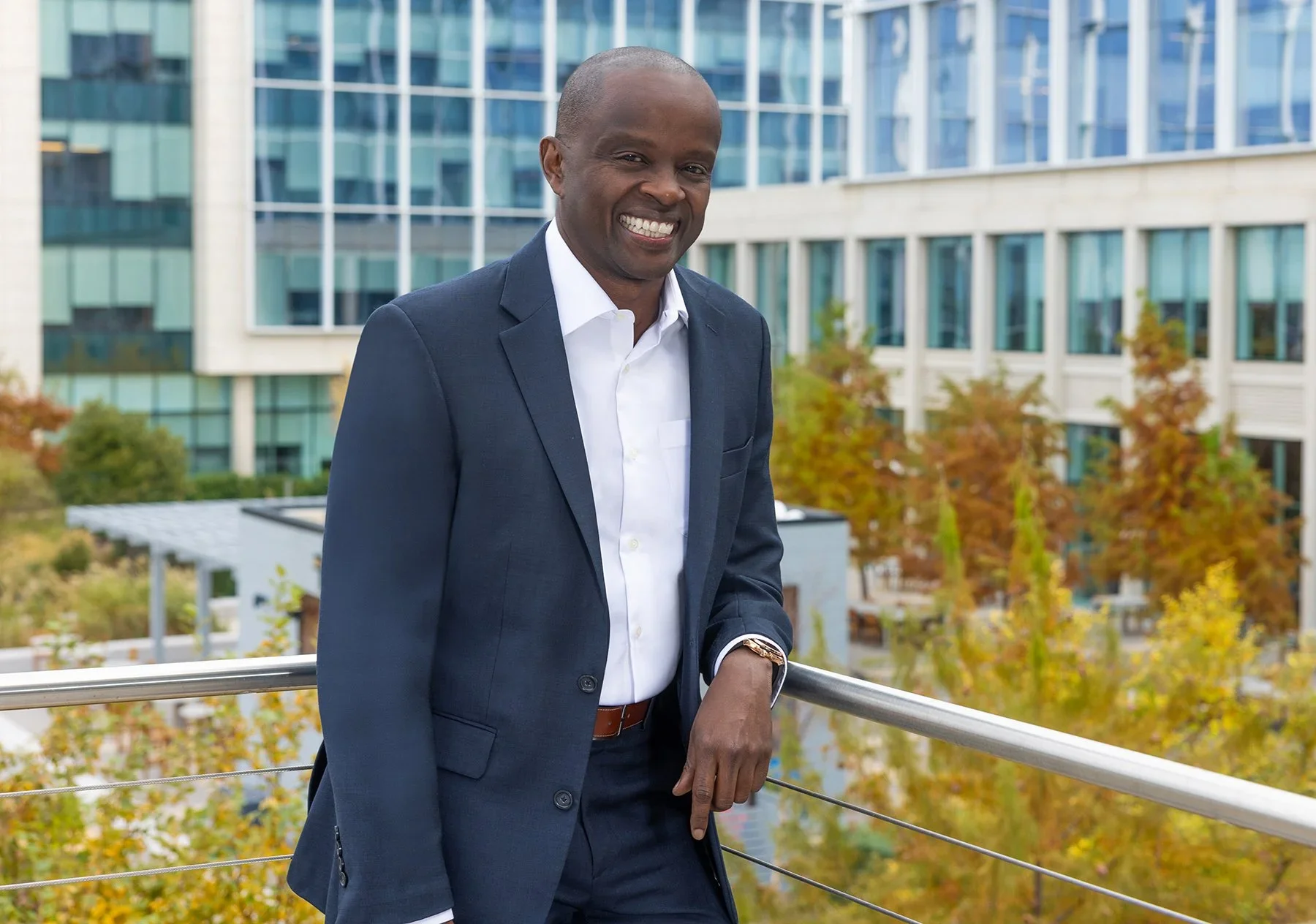 A smiling man in a navy suit and white shirt stands on a balcony with a metal railing, with modern glass buildings and trees with fall foliage in the background.