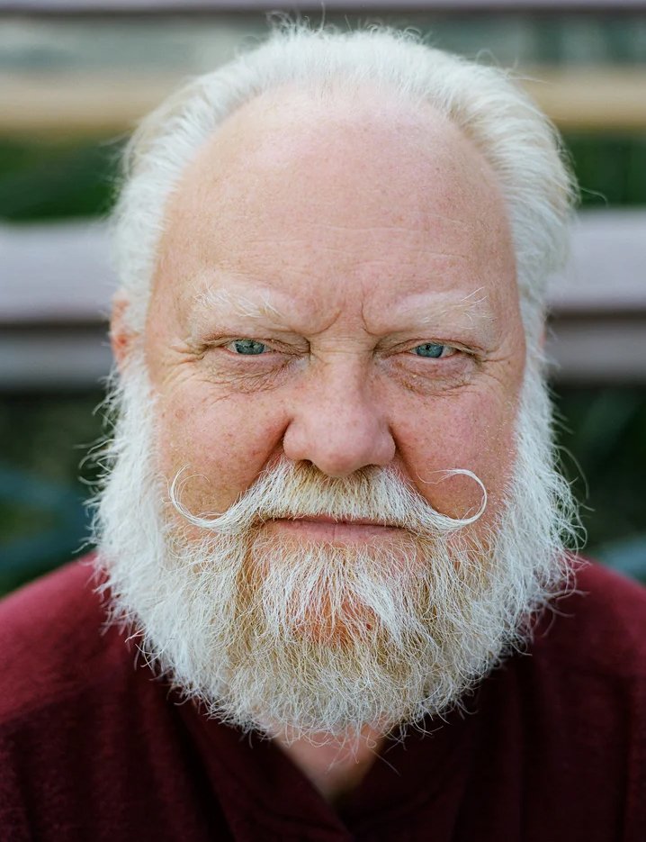 Close-up of an elderly man with white hair and beard, blue eyes, wearing a maroon shirt, outdoors with blurred greenery in the background.