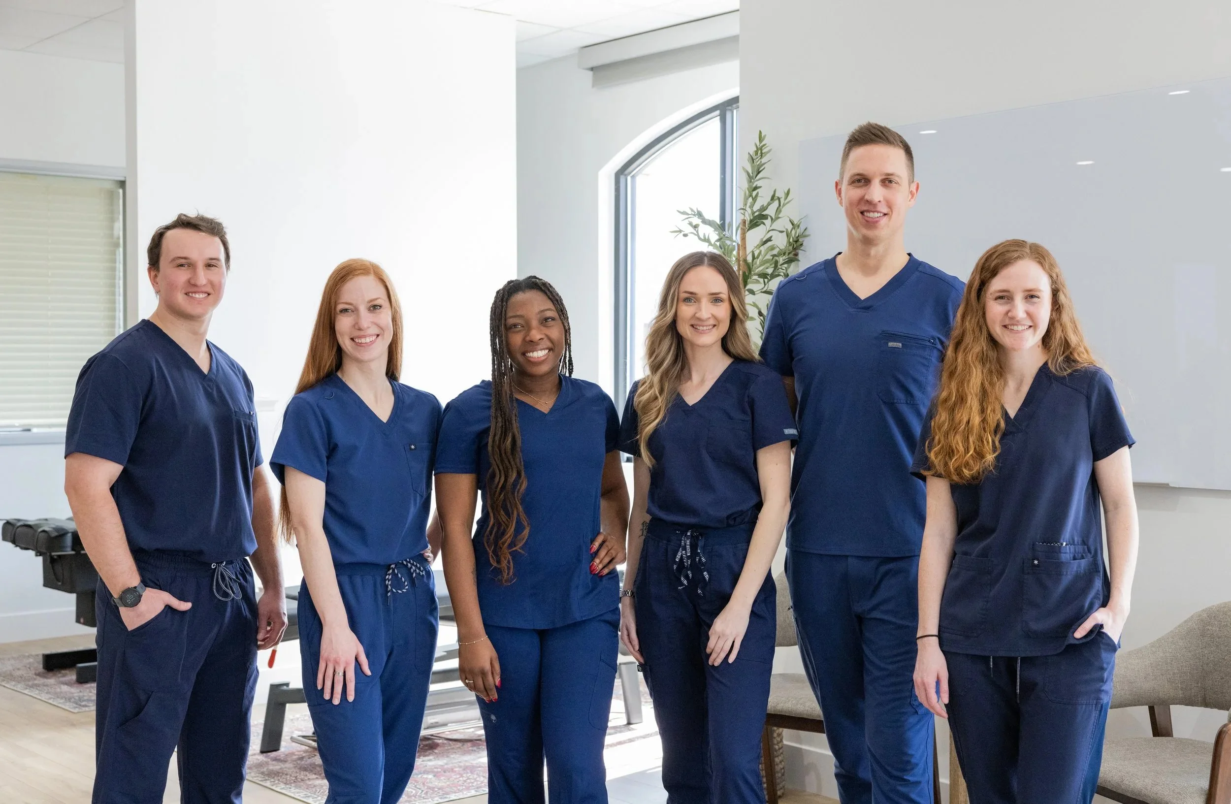 Group of six healthcare professionals wearing navy blue scrubs standing together in a modern medical office, smiling at the camera.