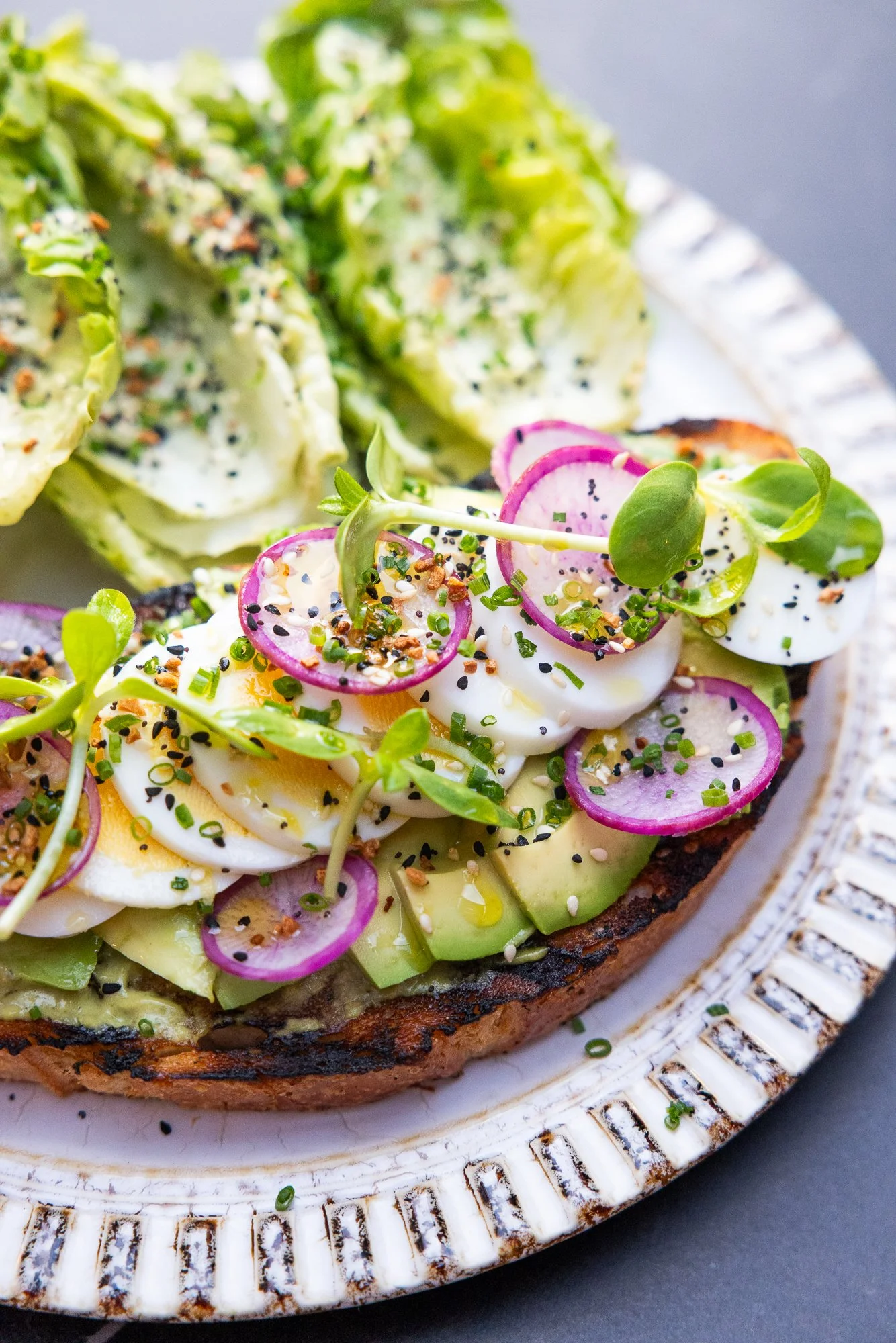 Close-up of avocado toast topped with soft-boiled eggs, radish slices, microgreens, and black sesame seeds, served on a decorative white plate.