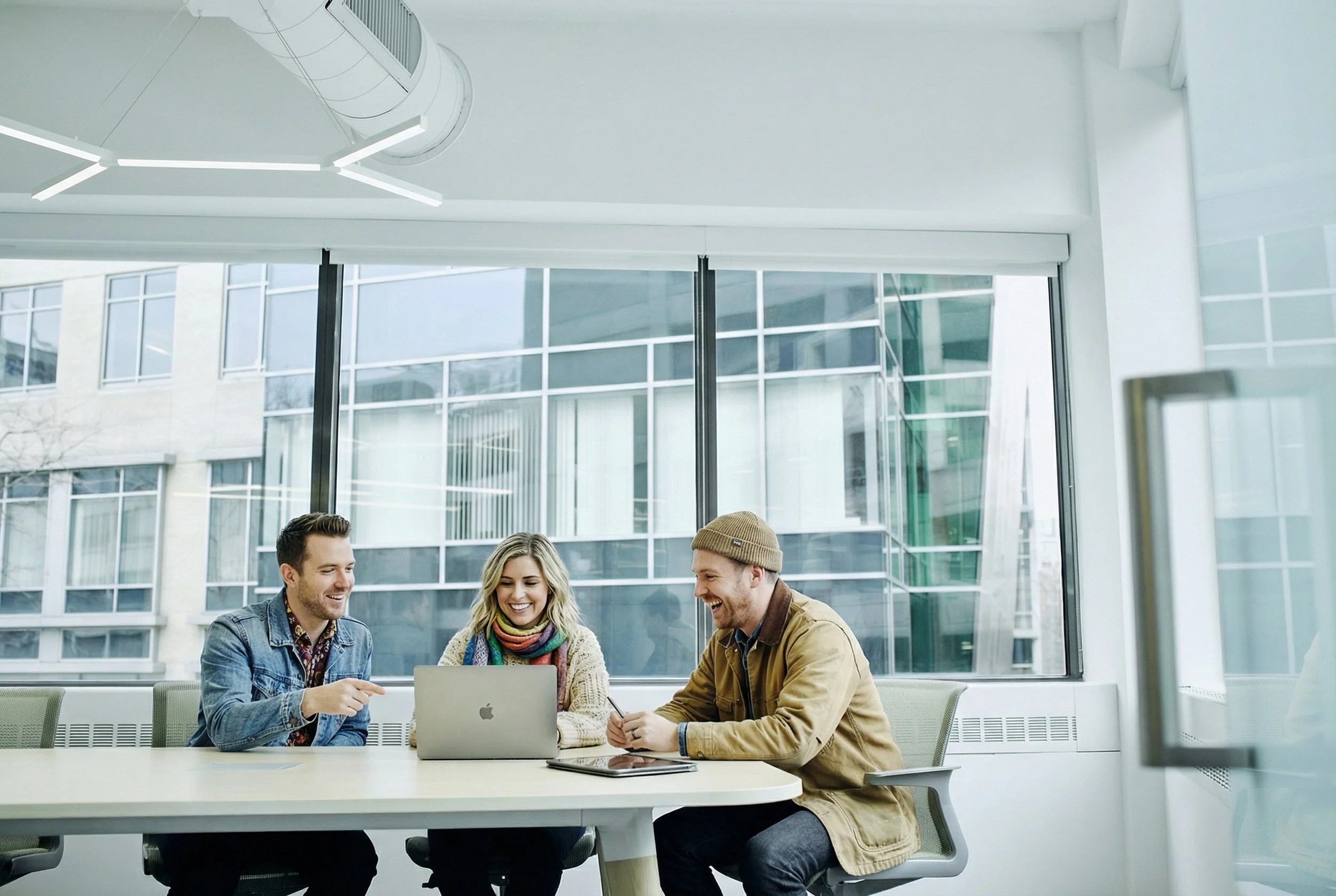 Three young adults sitting at a conference table in a modern office, smiling and talking while looking at a laptop.