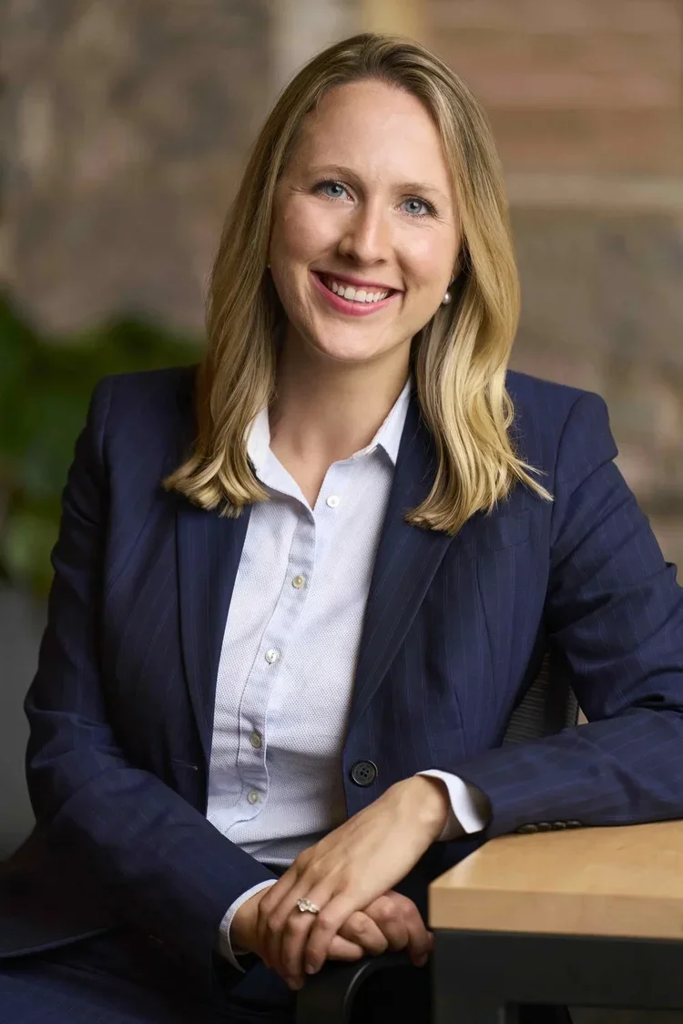 A smiling woman with blonde hair, wearing a navy blue blazer and a light blue shirt, sitting at a desk with her hands folded, in an office setting.