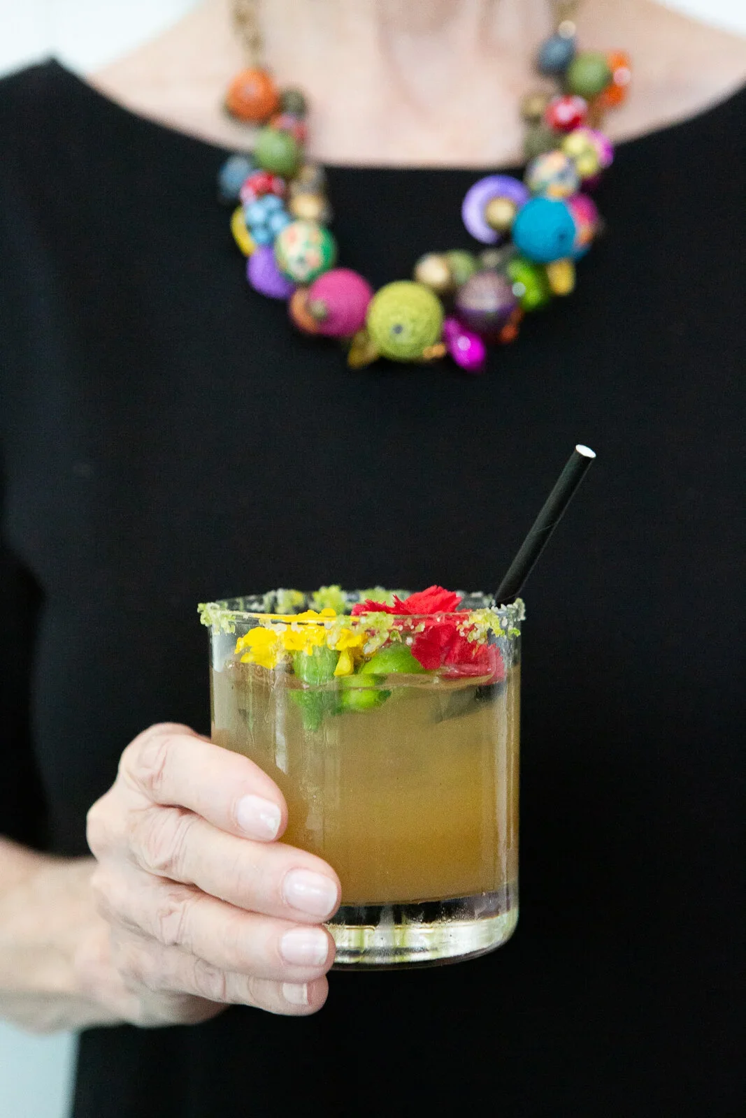 A person holding a glass of light brown cocktail garnished with colorful flower petals and a black straw.