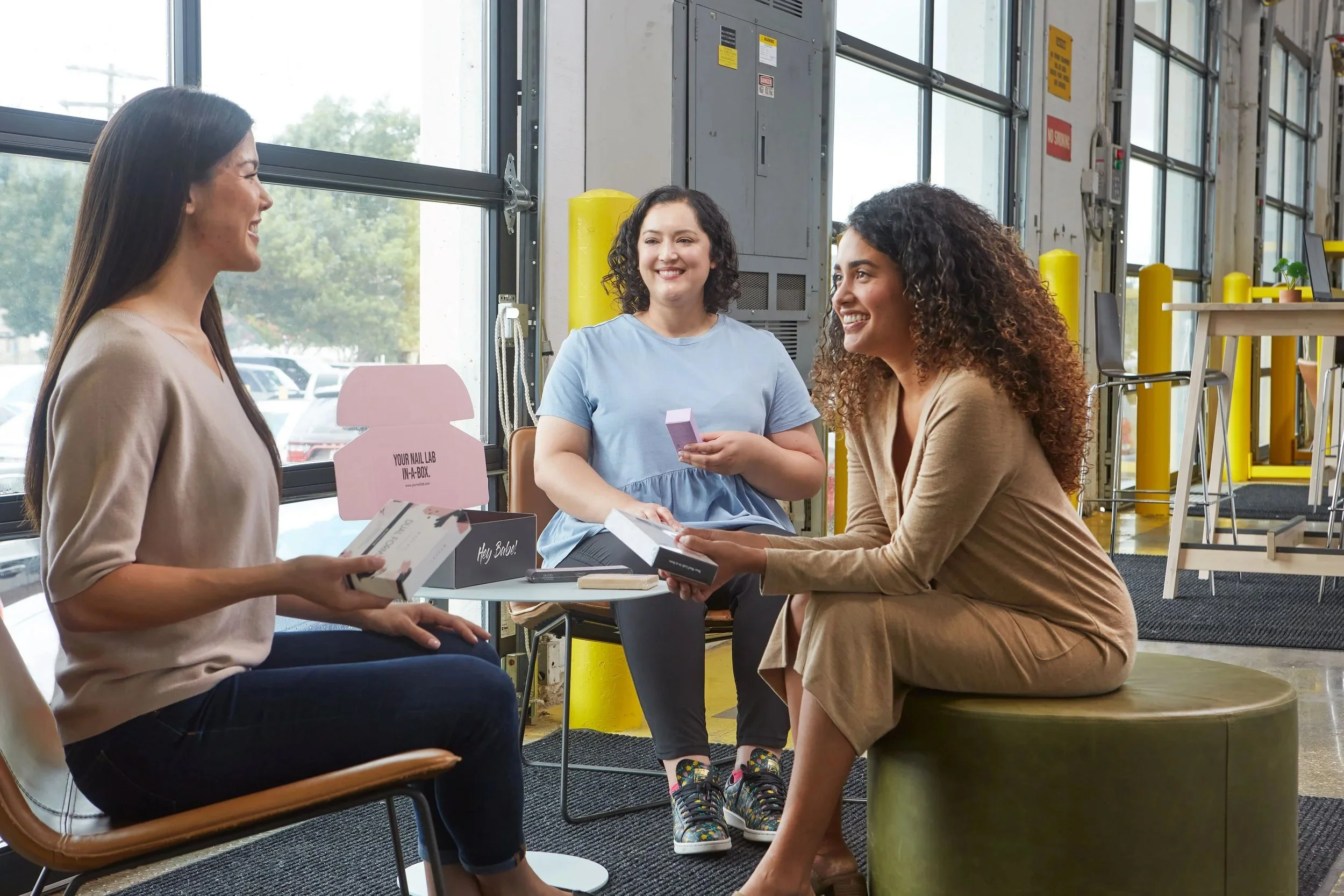 Three women talking inside a bright, industrial-style store or coffee shop, with items and large windows visible in the background.