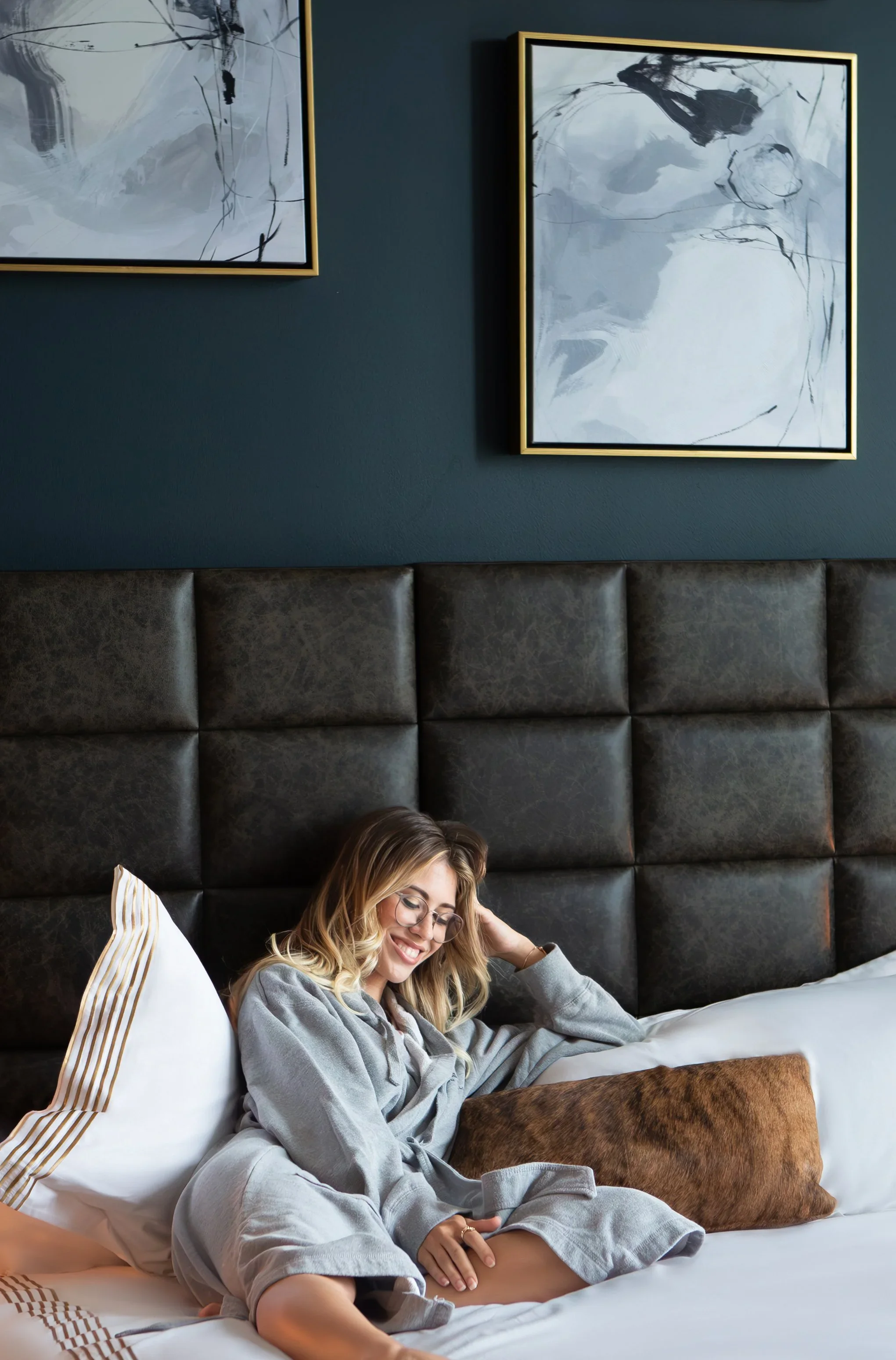 A woman with glasses wearing a gray robe, sitting on a bed and smiling, with tufted dark leather headboard, white and gold striped pillows, brown throw pillow, and abstract black and white artwork on a dark blue wall.