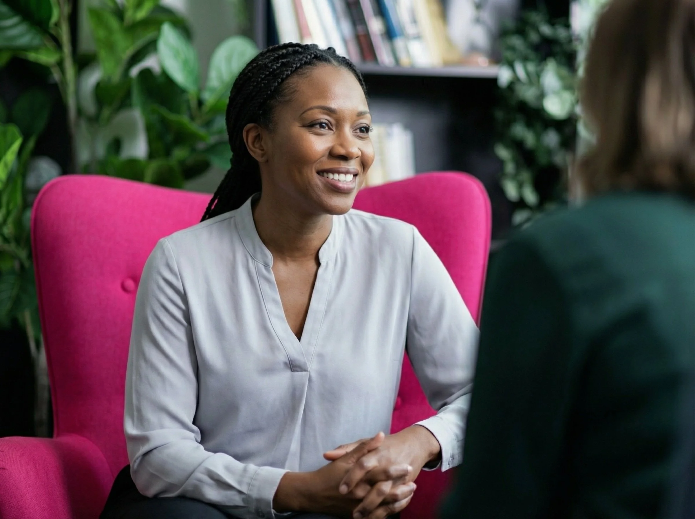A woman with braided hair smiling during a conversation in an office or lounge area with pink chairs and green plants in the background.