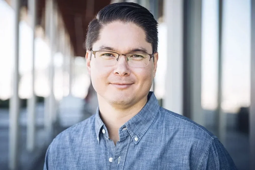 Close-up portrait of a man with glasses and dark hair, wearing a blue button-up shirt, standing in a modern building with large windows.