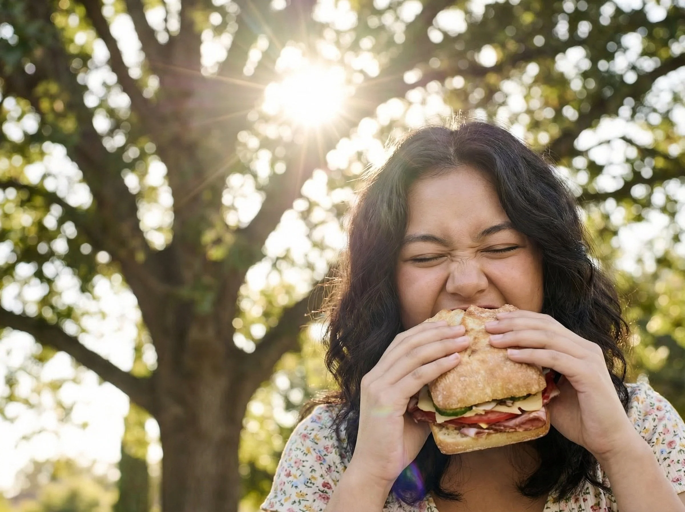 A woman with curly dark hair is taking a big bite of a sandwich outdoors in a park, with sunlight shining through trees in the background.