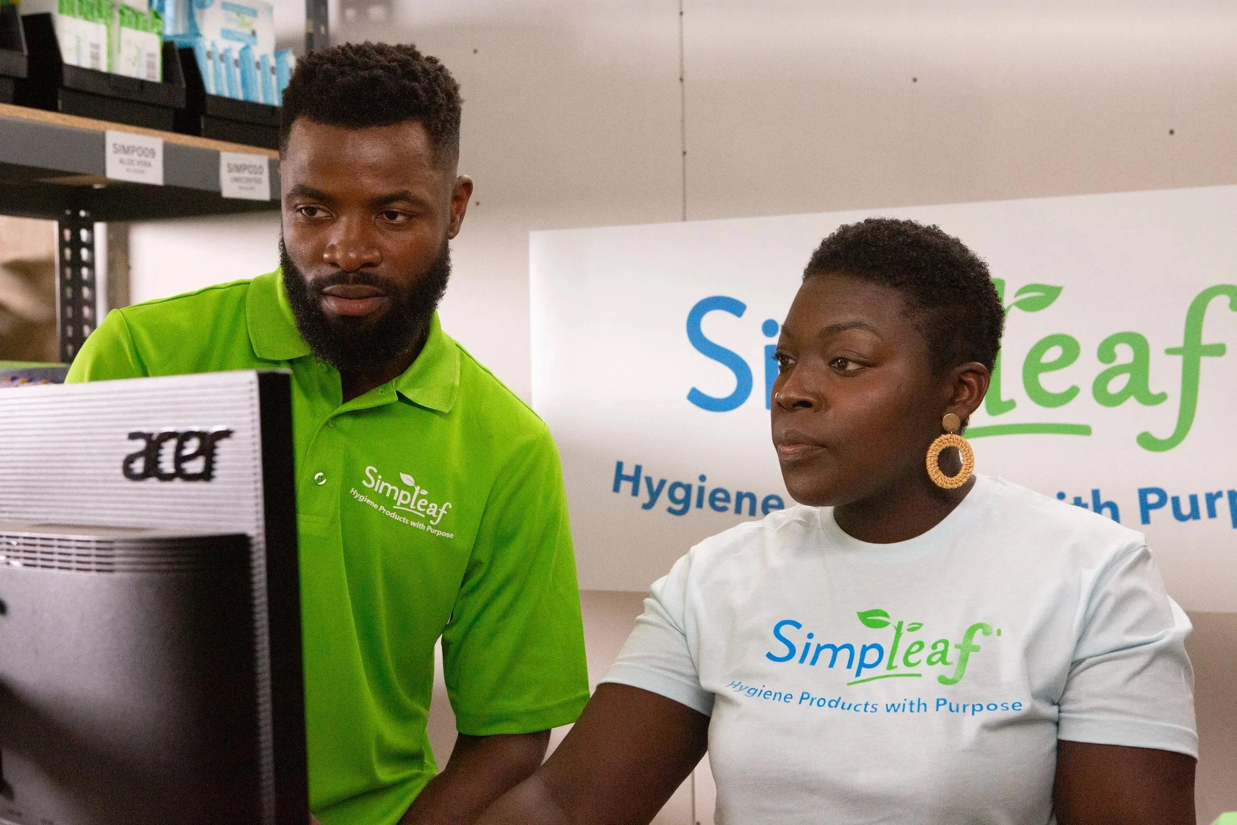 A man and woman at a workspace, both wearing shirts with the 'Simpleaf' logo and the tagline 'Hygiene Products with Purpose'. The man is wearing a bright green polo shirt, and the woman is wearing a white T-shirt. They are looking at a computer monit