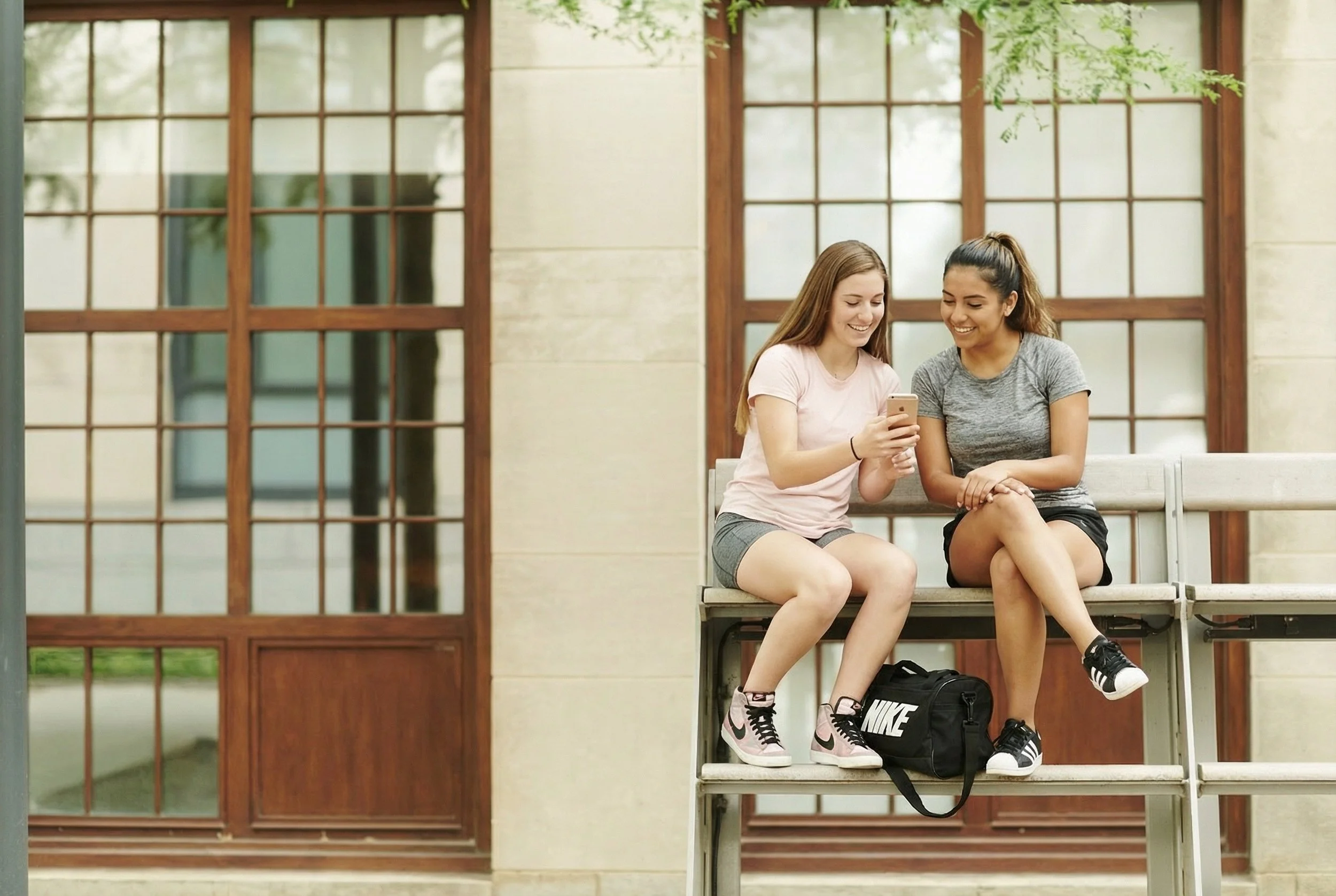 Two young women sitting on a bench outdoors, looking at a smartphone together, smiling, with a building and large windows behind them.