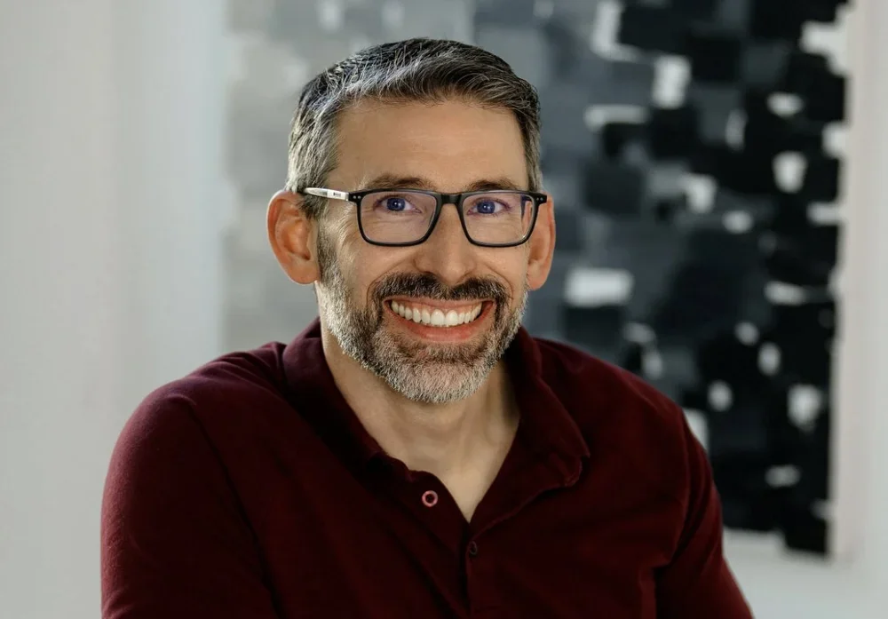 Smiling middle-aged man with glasses, short dark hair, and a beard with gray, wearing a burgundy shirt in an indoor setting.