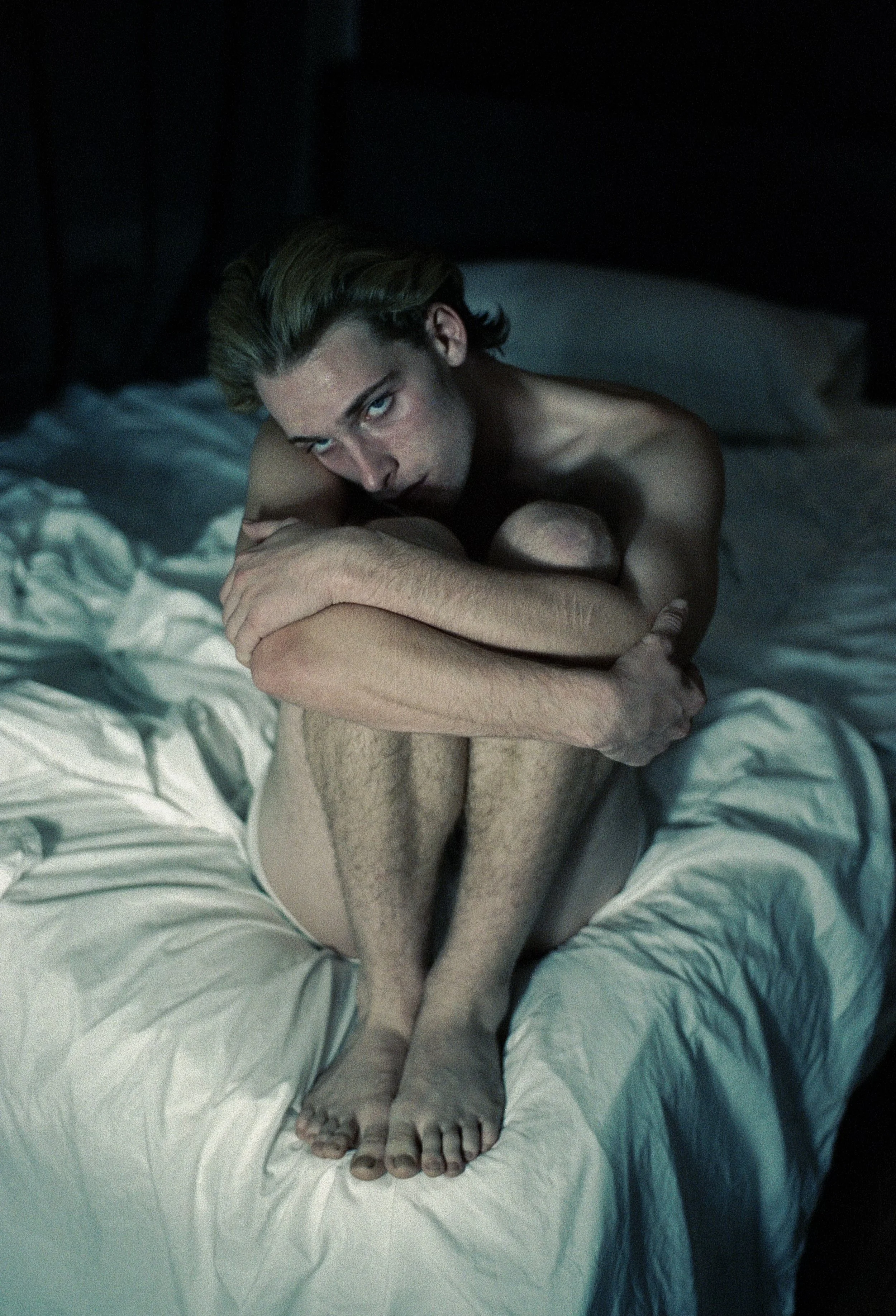 A young man with light skin and blonde hair sitting on a bed with white sheets, hugging his knees, looking directly at the camera with a serious expression, in a dimly lit room.