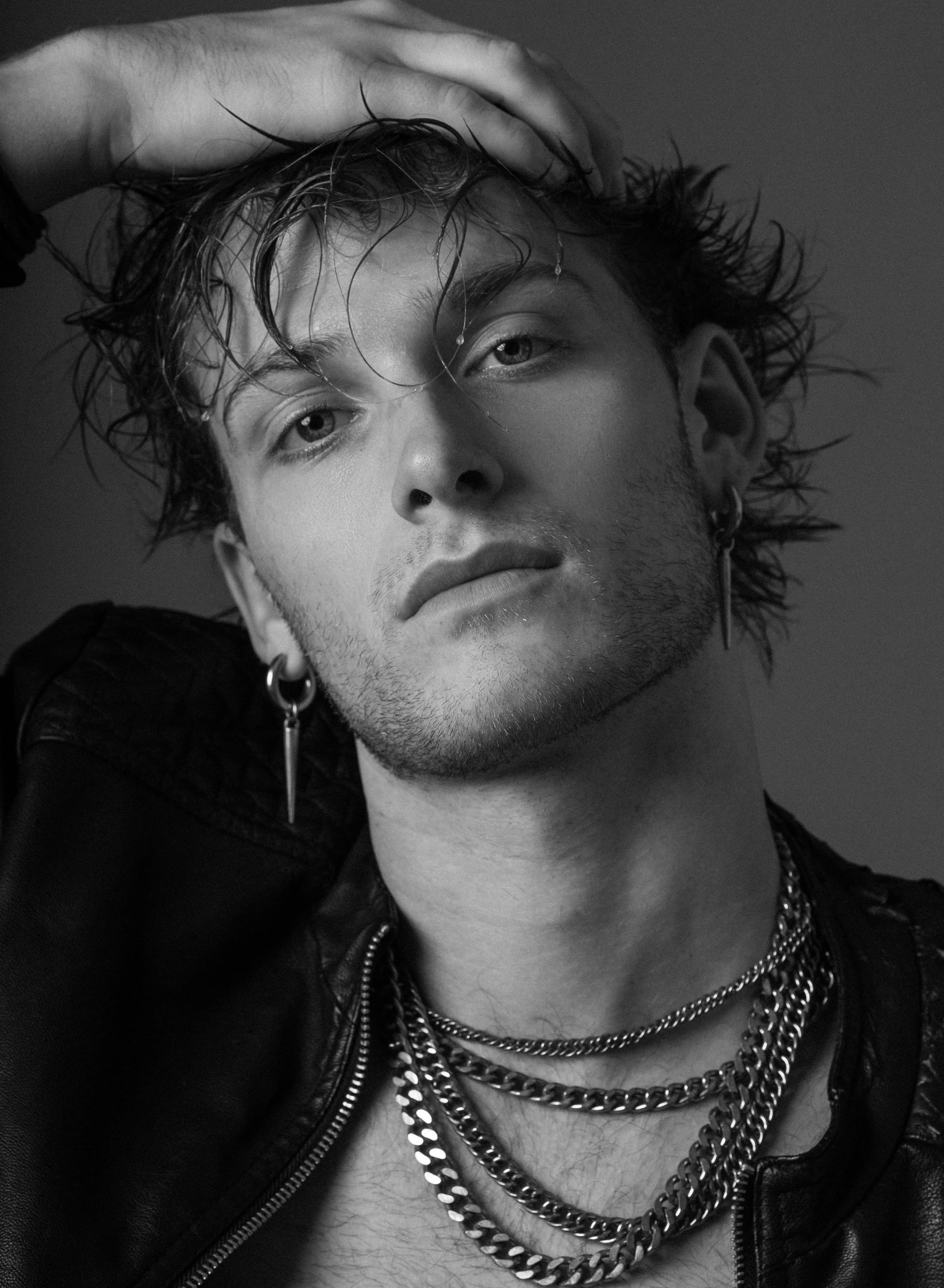 Black and white portrait of a young man with wet hair, multiple necklaces, leather jacket, and earrings, posing with one hand on his head.