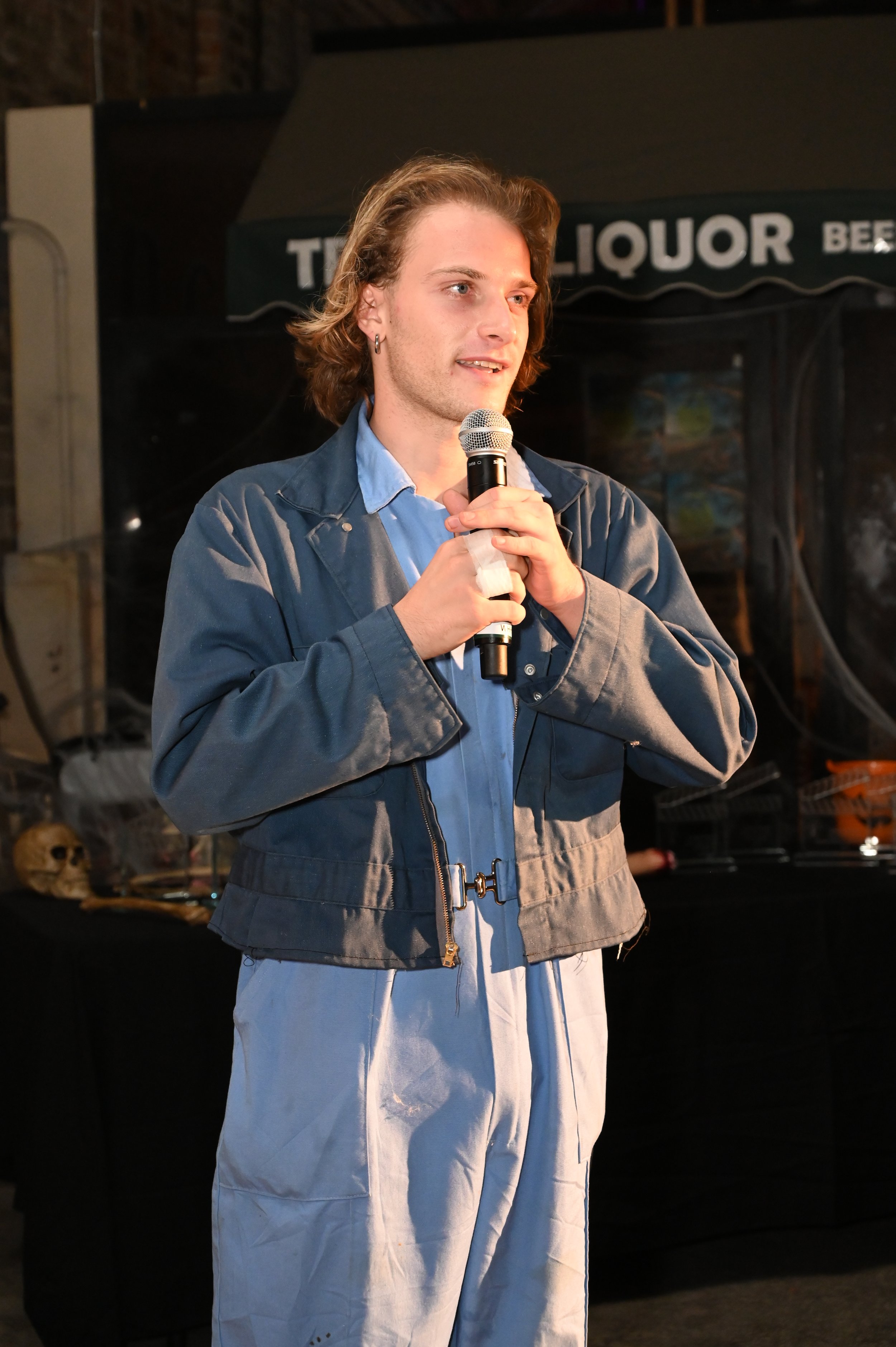 A young man with shoulder-length hair wearing a blue shirt and gray jacket holding a microphone, speaking at an event with a black booth behind him marked 'LIQUOR'.