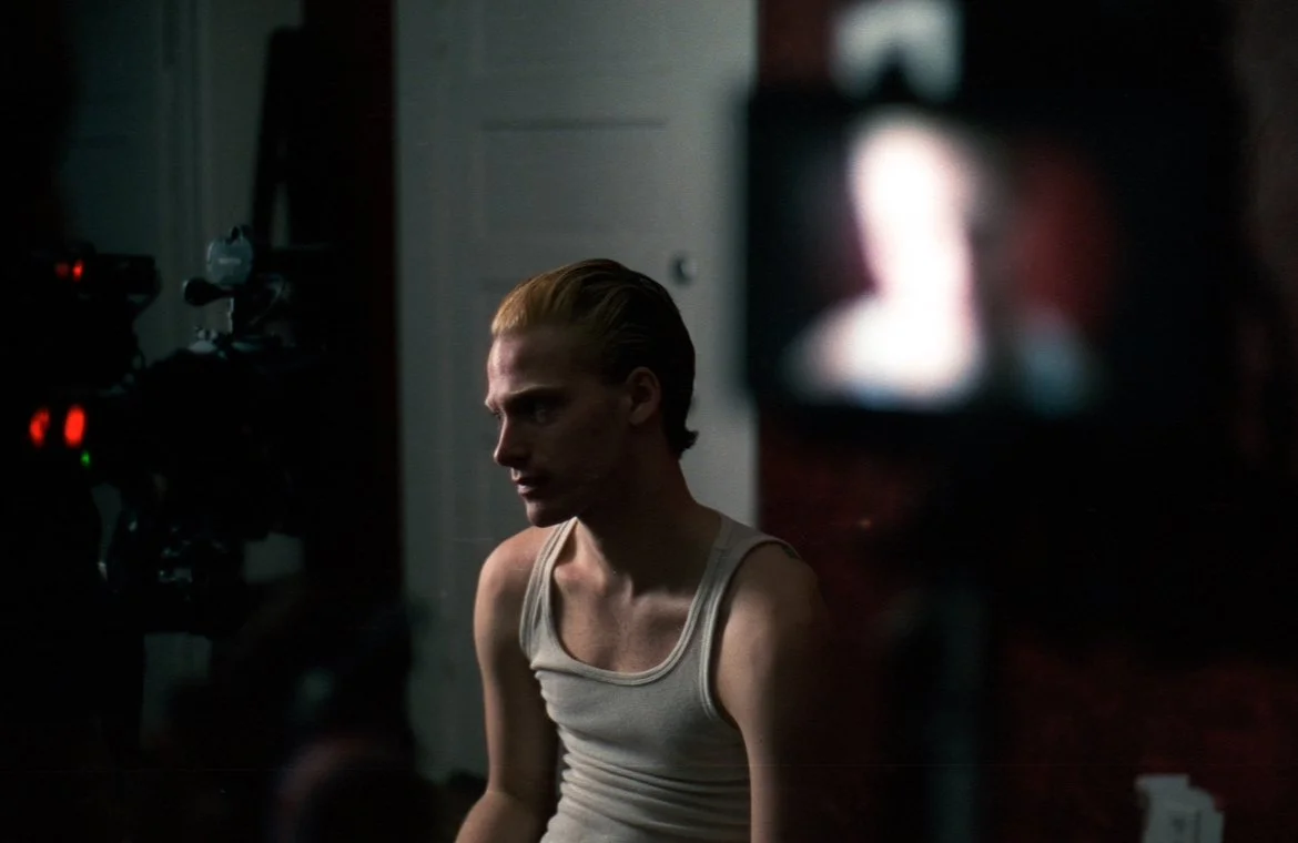 A man with slicked-back hair wearing a white tank top, sitting in a dimly lit room with a serious expression, with a camera on the left and a blurred screen on the right.