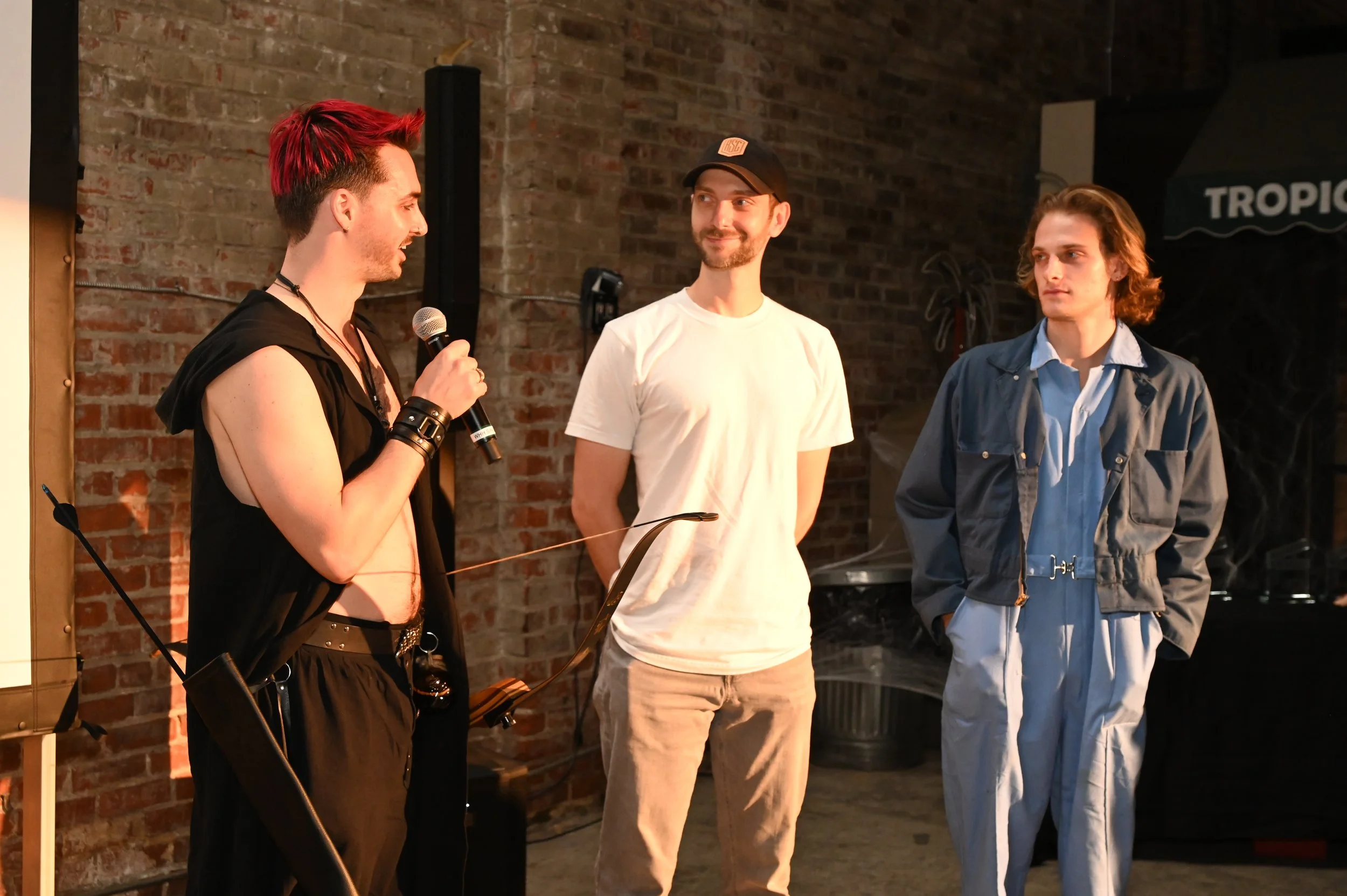 Three young men standing indoors against a brick wall, two listening to one who is speaking into a microphone. One man has bright red hair, wearing a sleeveless hoodie and bracelets. Another has brown hair, wearing a white t-shirt and a black cap, smiling. The third has wavy brown hair, wearing a blue jacket and light-colored pants.
