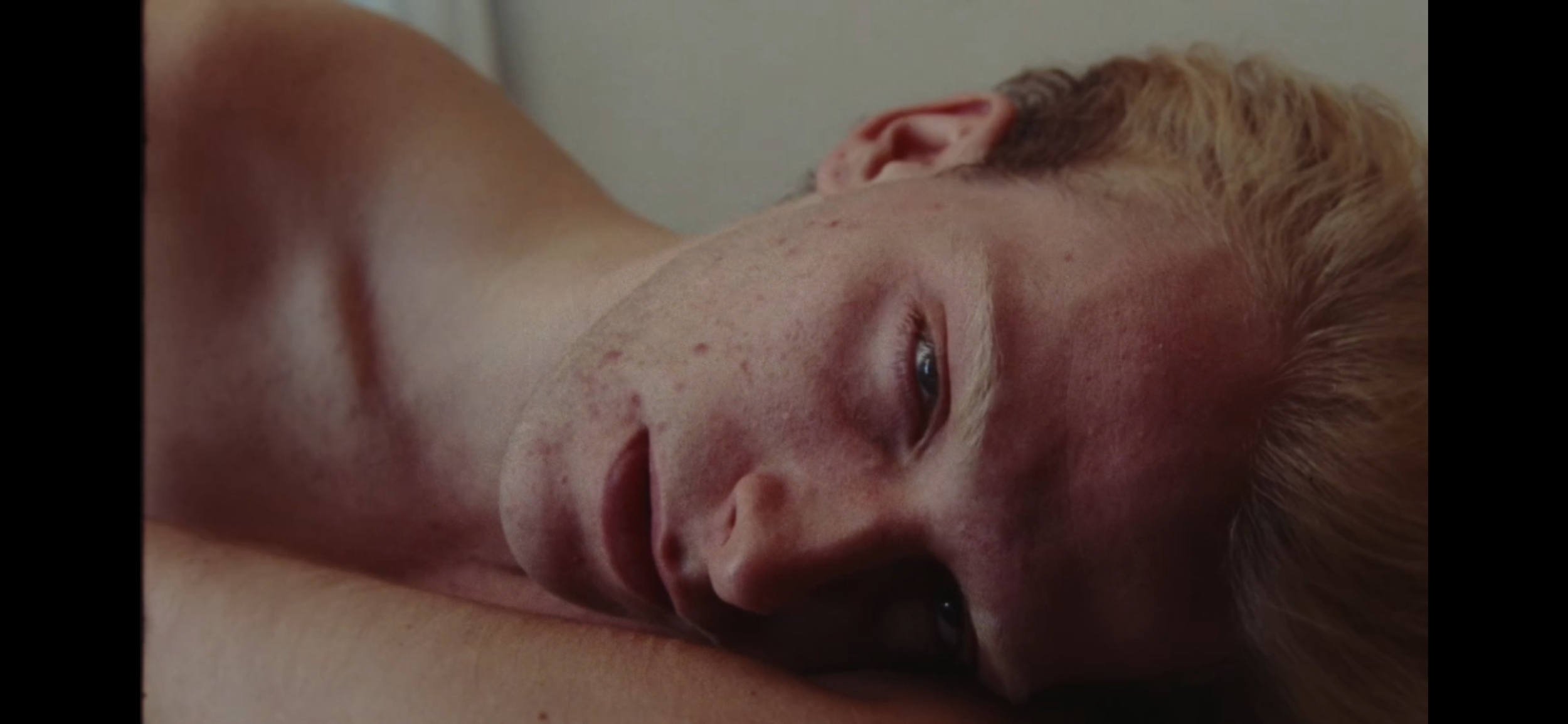 A young man with light skin and blond hair is lying down with his head resting on his arm, looking into the camera.