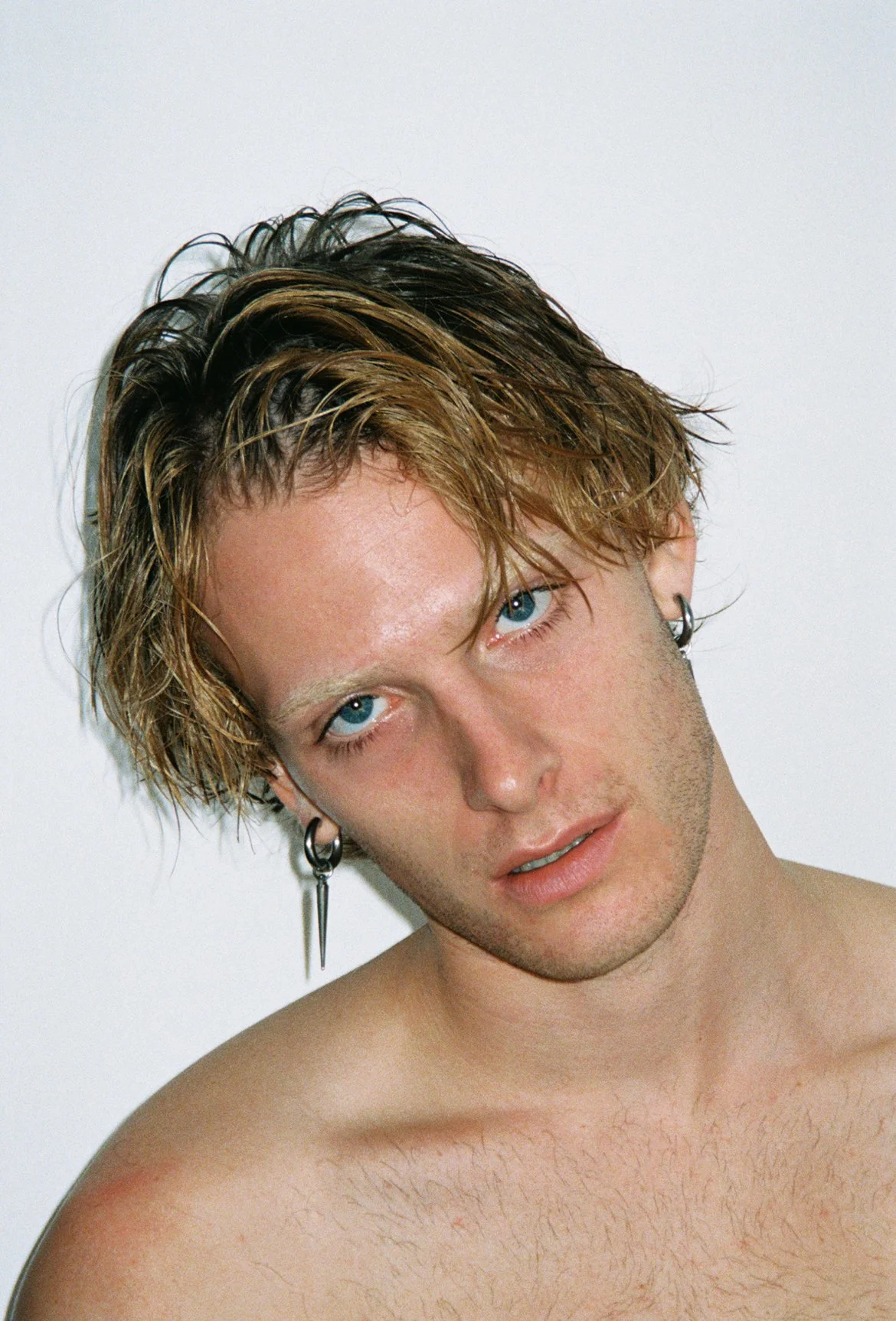 A young man with light brown, wet hair, blue eyes, and earrings poses shirtless in front of a plain background.