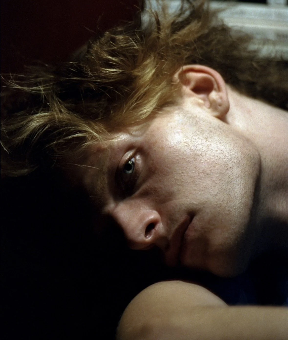 A close-up of a person's face with short, curly red hair, lying on the ground, looking towards the camera with a serious expression.