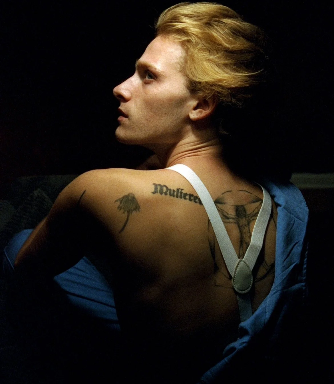 Side profile of a person with blonde hair, tattoos on their shoulder and back, wearing a blue shirt and white suspenders, against a dark background.