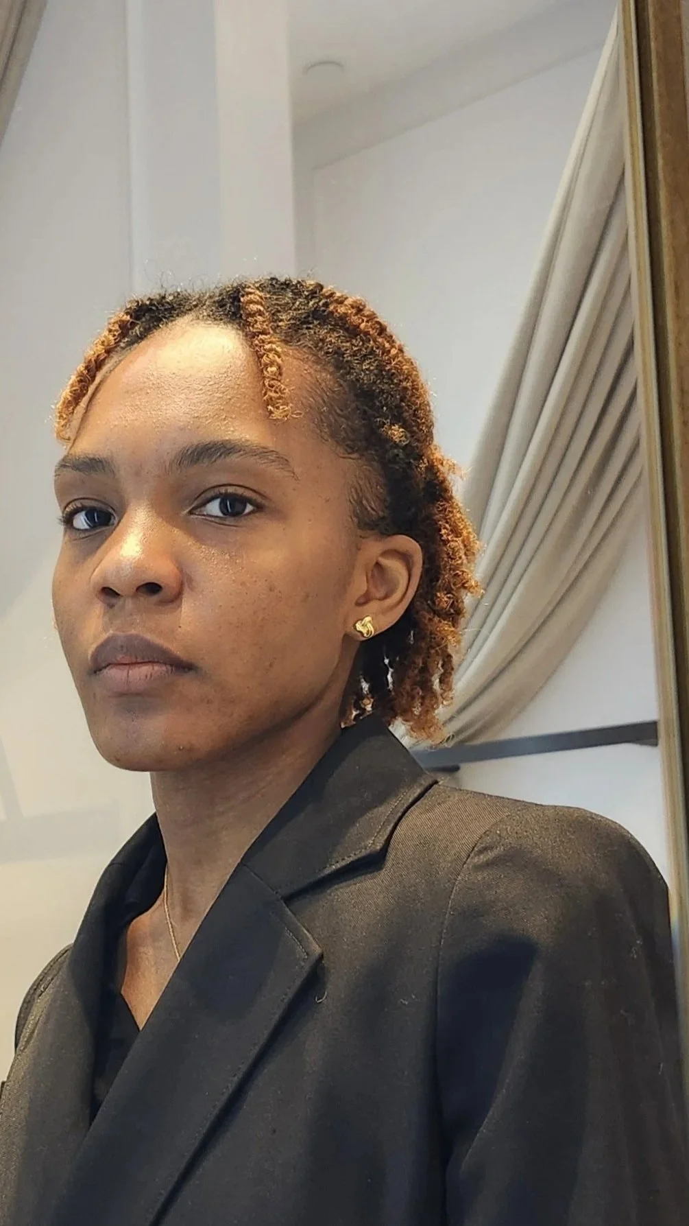 A person with short curly hair, wearing gold earrings, dressed in a black blazer, looking into a mirror in a room with beige curtains.