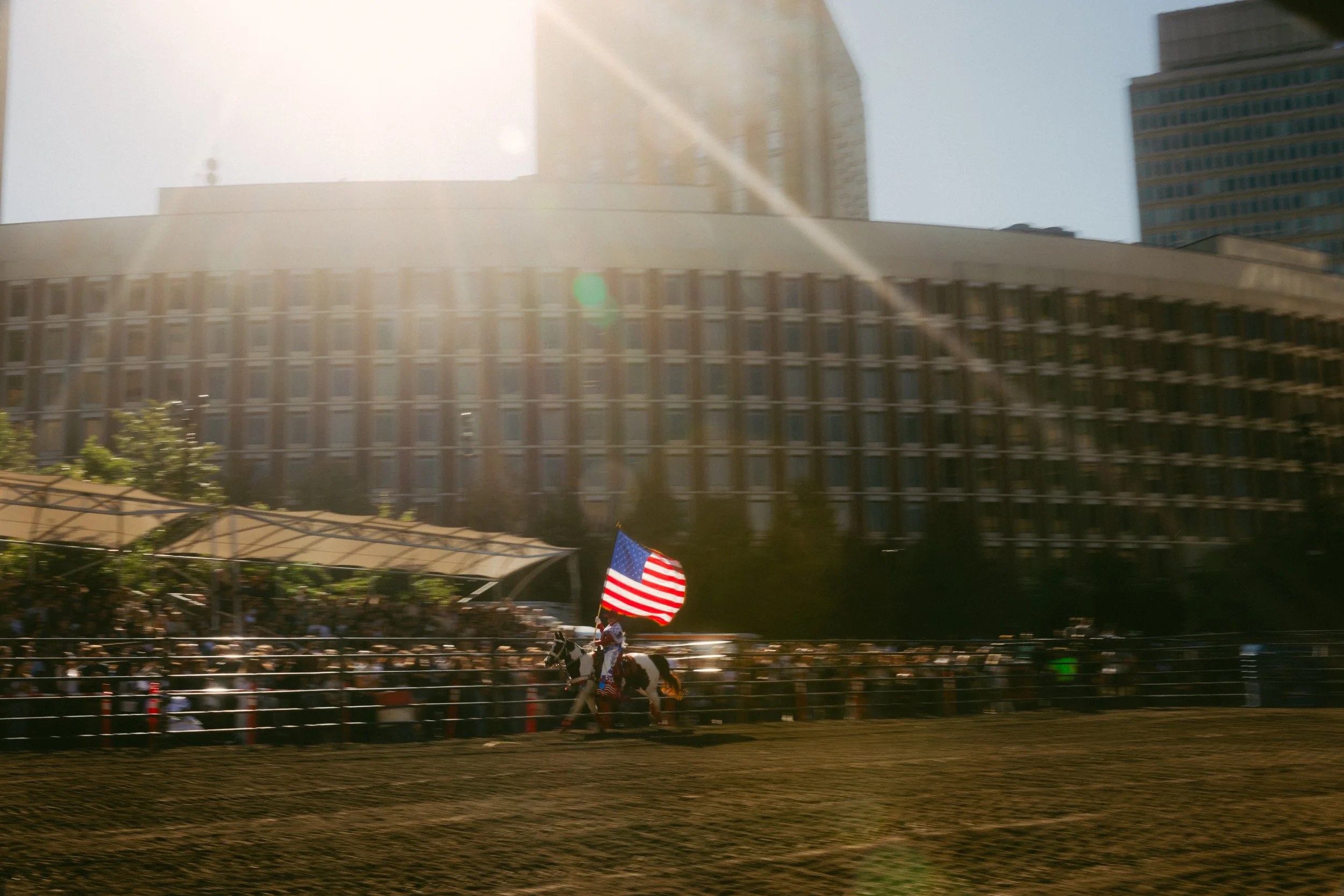 Horse rider carrying an American flag at a rodeo event, with a large crowd and modern buildings in the background, and sunlight creating lens flare.