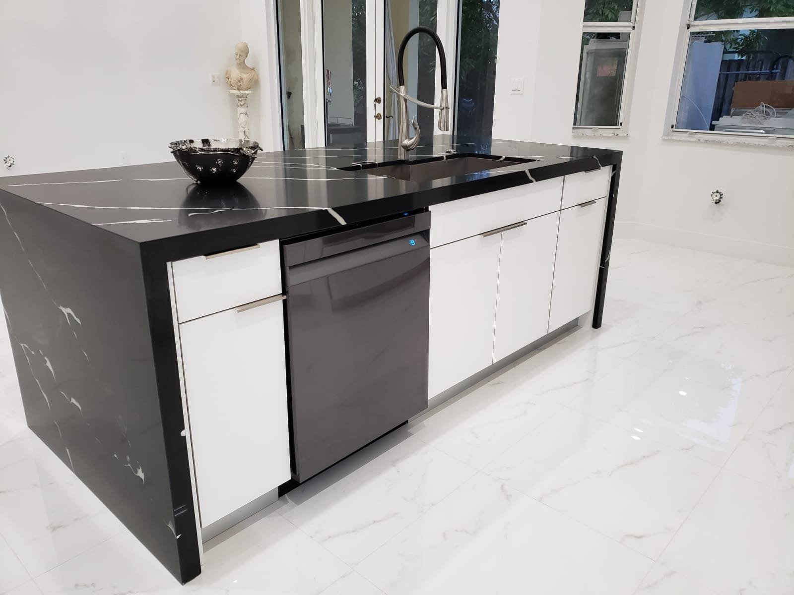 Modern kitchen island with a black countertop, white cabinets, a built-in black dishwasher, a black bowl, and a black faucet in front of large windows and sliding doors.