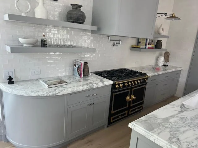 Modern kitchen with gray cabinets, marble countertops, and white tiled backsplash. Open shelves hold white and black decorative items, glassware, and books. A black and gold oven is centered between the cabinets.