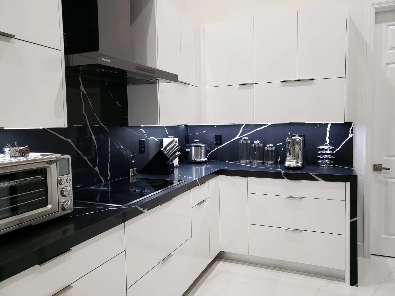 Modern kitchen with white cabinets, black marble countertops, and a black marble backsplash with white veining. Small appliances include a toaster oven, kettle, and containers.
