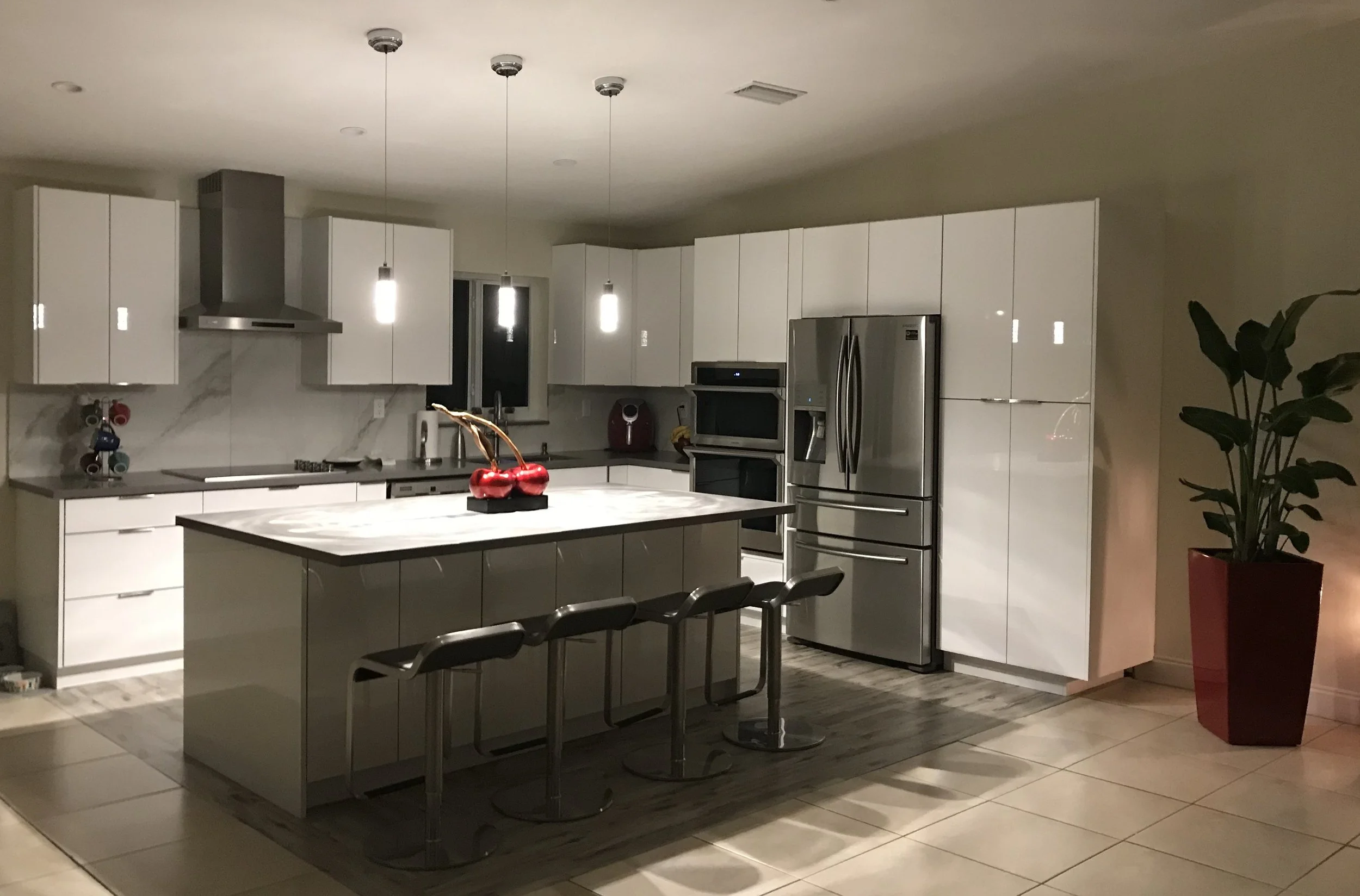Modern kitchen with white cabinets, stainless steel refrigerator, built-in oven, and marble island with three chairs. Pendant lights hang above the island, and there is a potted plant in a red pot on the right side.